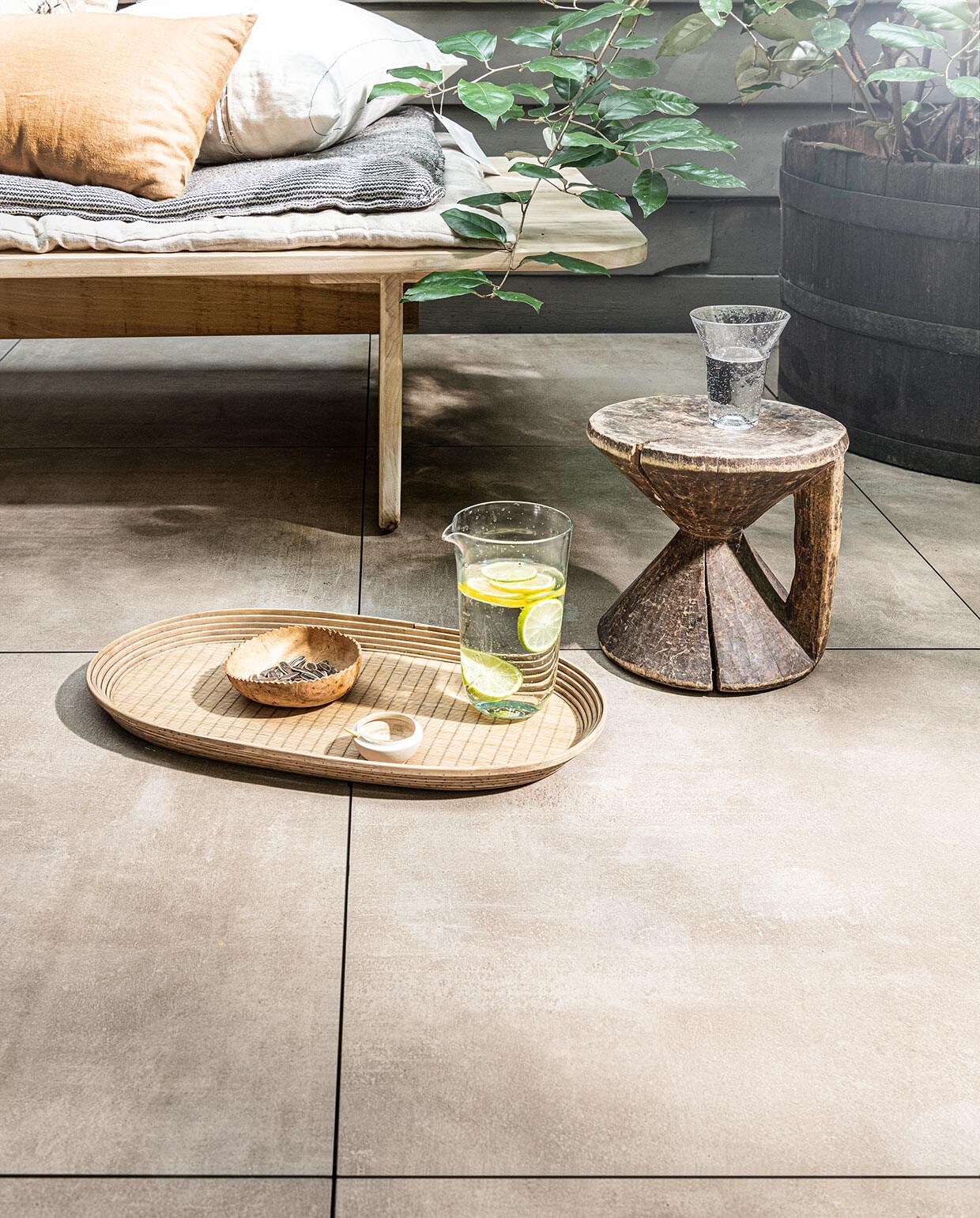 Tegel Solostone Uni Beton Olive, , 70x70x3,2 cm (lxbxh) (ook verkrijgbaar in Grey, Antracite en Taupe). , M.m.v. vtwonen.nl (jute bloempot van Vase the World, aardewerken bloempot van Serax, glazen karaf van Serax, glas van Serax), sukha.nl (houten daybed, groot kussen Island Friend), iblaursen.nl (dienblad, gestreept matraskussen). Overig privébezit. vtwonen 03-2021 | Productie Tanja Saarberg | Styling Kim van Rossenberg | Fotografie Sjoerd Eickmans