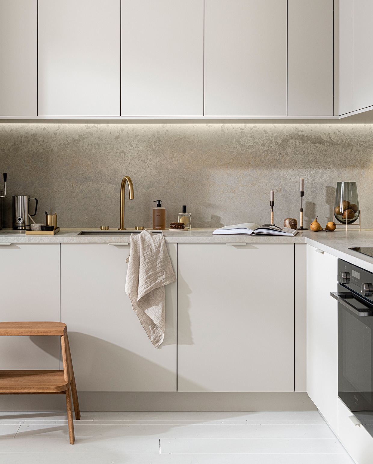 Keuken Basic Greige, vanaf € 21.500,-,  M.m.v. thefinestore.com (houten krukje van Hay, messing dienblad en beker van Ferm Living, bronzed brass kandelaren Duca van Menu), flinders.nl (vaas van gerookt glas op messing pootjes van Menu), kikubari.nl (spray van Lingua Planta), vtwonen.nl (theedoek uit de vtwonen collectie), skins.nl (handzeep Suede Byredo), sjostrandcoffee.nl (melkschuimer). Overig privébezit. vtwonen 03-2021 | productie Tanja Saarberg | styling Kim van Rossenberg | fotografie Sjoerd Eickmans