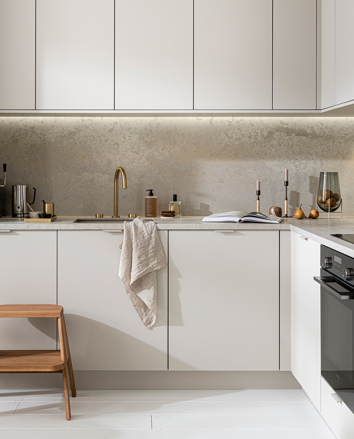 Keuken Basic Greige, vanaf € 21.500,-,  M.m.v. thefinestore.com (houten krukje van Hay, messing dienblad en beker van Ferm Living, bronzed brass kandelaren Duca van Menu), flinders.nl (vaas van gerookt glas op messing pootjes van Menu), kikubari.nl (spray van Lingua Planta), vtwonen.nl (theedoek uit de vtwonen collectie), skins.nl (handzeep Suede Byredo), sjostrandcoffee.nl (melkschuimer). Overig privébezit. vtwonen 03-2021 | Productie Tanja Saarberg | Styling Kim van Rossenberg | Fotografie Sjoerd Eickmans