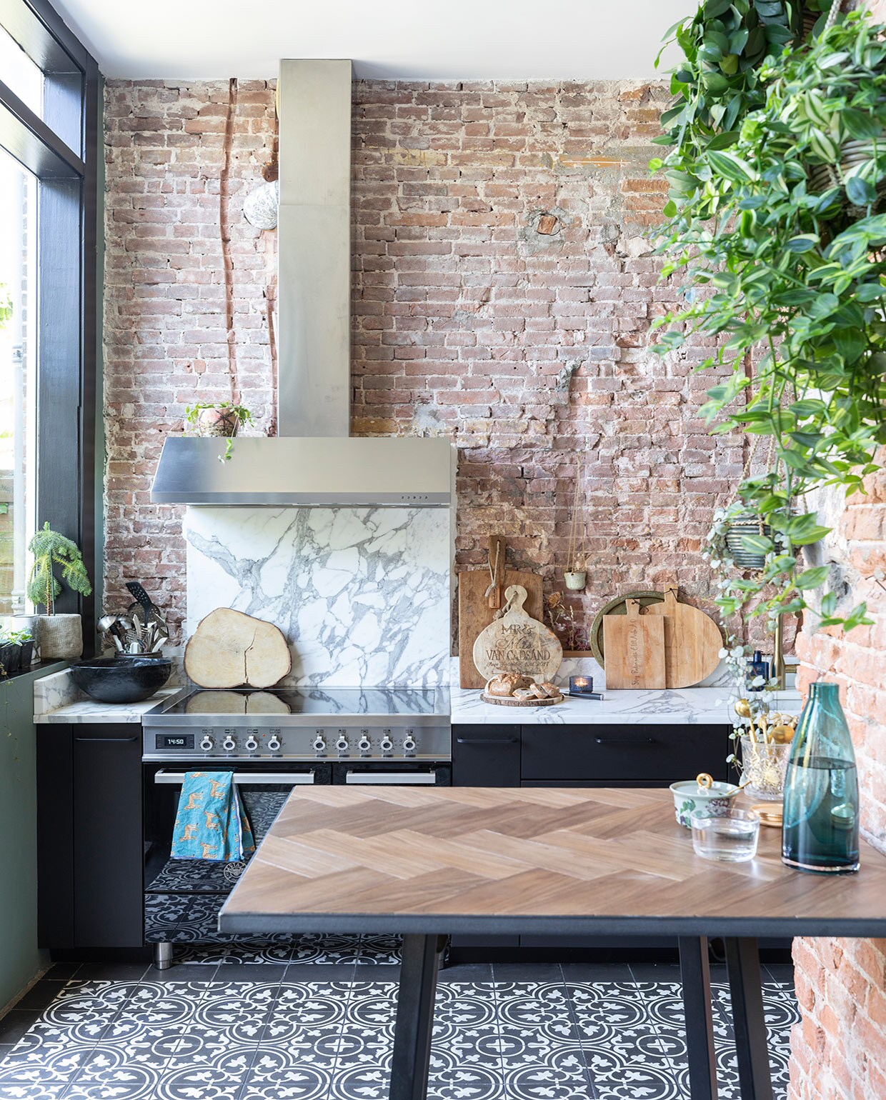 Portugese vloertegels Designtegels, keukenkastjes Ikea, werkblad en spatwand van Carrara marmer door Swaalf uit Haarlem, fornuis Bertazzoni, groene muurverf Green Smoke en zwarte verf kozijnen Pitch Black Farrow &amp; Ball, pot in vensterbank, blauwe waterkan, waterglas en zwarte schaal Ikea, snijplank boomstam laten maken op Bali, snijplanken gekregen van vrienden, hangpot Dekatuin, pot op afzuigkap gekregen, hoge bartafel onderstel van Hay met massief eikenhouten blad gemaakt door SkillsXL, suikerpot van Essenza, vaas onbekend. vtwonen 01-2021 | productie en fotografie Margriet Hoekstra