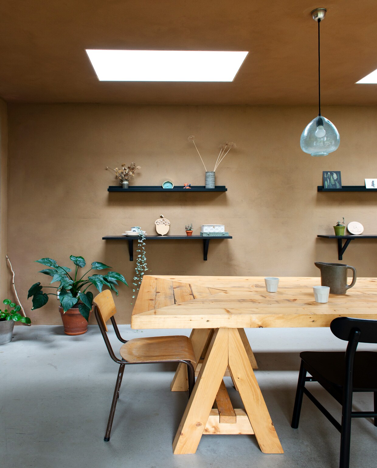Wanden en plafond van onbehandeld leemstuc, vloer van beton. Muurplanken, vloerkleden, stoelen , hanglampen . Tafel en bank gemaakt door Joost. Vintage schoolstoel, schenkkan en vazen kringloop vtwonen 01-2021 | styling Esther Schuivens | fotografie Caroline Coehorst | tekst Chris Muyres