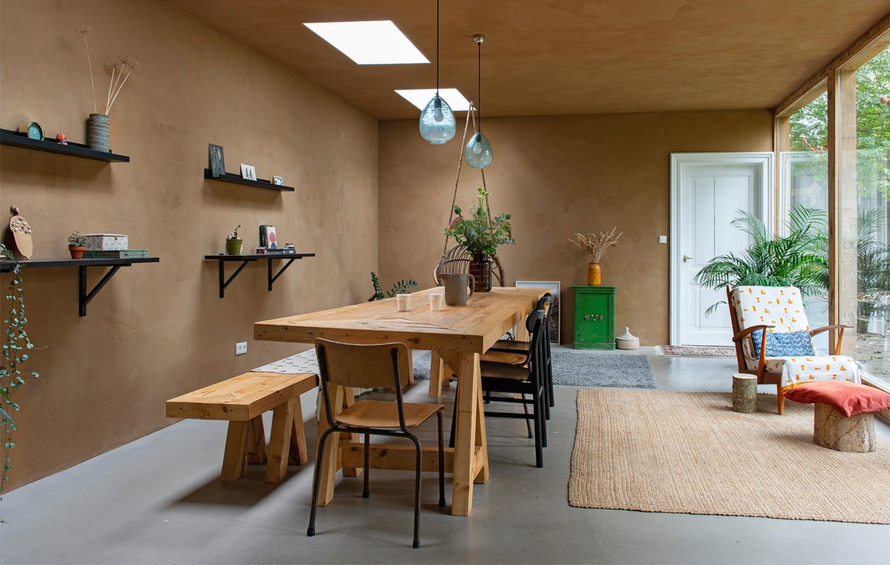 Wanden en plafond van onbehandeld leemstuc, vloer van beton. Muurplanken, vloerkleden, stoelen Ikea, hanglampen Sissy-Boy. Tafel en bank gemaakt door Joost. Vintage schoolstoel, schenkkan en vazen kringloop, kluis en fauteuil vintage vondsten. Rieten mand Wereldwinkel. Deurmat vintage, deken en kussen op fauteuil Ted &amp; Tone. Rood kussen Dille &amp; Kamille. Bijzettafel en kruk zelfgemaakt van boomstammen uit de tuin. vtwonen 01-2021 | Styling Esther Schuivens | Fotografie Caroline Coehorst