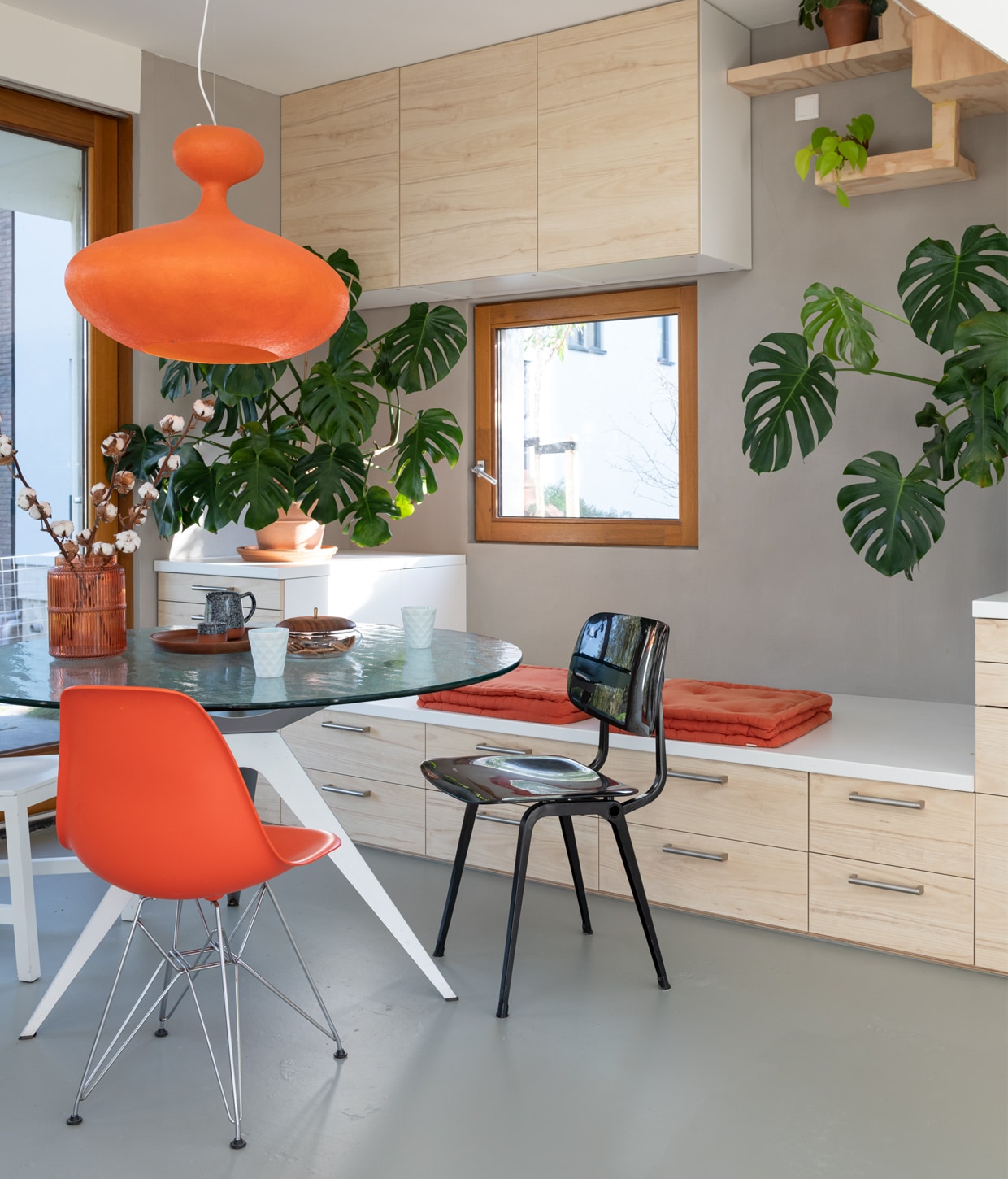 Zandcementdekvloer met vloercoating, trap en houten muren van vurenhout en radiplex, eettafel Ben Hoek Meubelarchitectuur, lamp Kundalini E.T.A. Sat, oranje stoel Charles Ray Eames, zwarte stoel Revolt van Friso Kramer, witte stoel Joep van Lieshout, oranje vaas Kwantum, dienblad H&amp;M Home, kan Søstrene Grene, lichtblauwe mokken kringloop, koektrommel met houten deksel en grijze aardewerken pot vintage, oranje palletkussens tuincentrum, terracotta potten Intratuin en Ikea, handgrepen lades Ikea. vtwonen 01-2021 | Productie en fotografie Caroline Martinot | Styling Sharon Roest