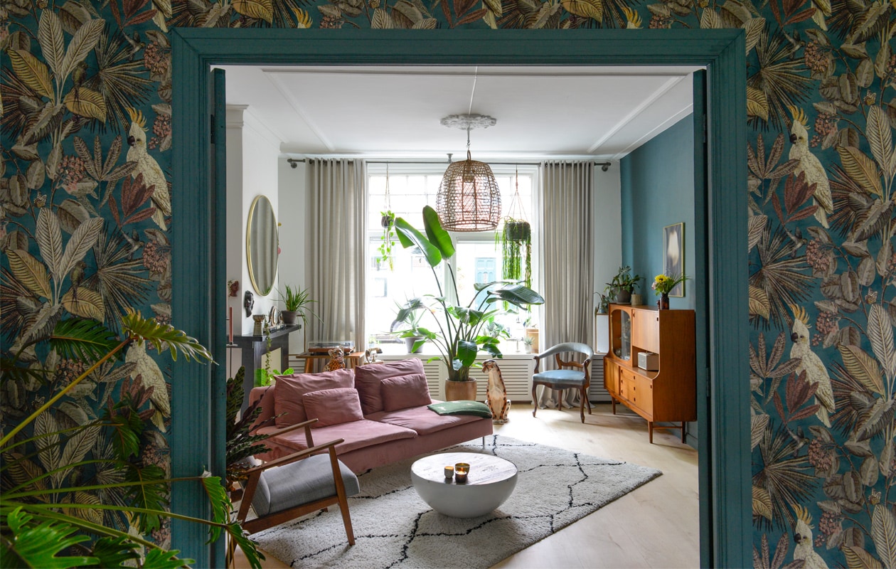 Bank en grijze fauteuil Ikea, bankhoes Norsemaison. Groene plaid, waxinehouders en spiegel Sissy-Boy. Salontafel veiling. Plafondlamp De Handelsfabriek. Kleed Karwei. Platenspelermeubel, vaas op schouw, blauwe stoel en kast vintage. Tijger en cheeta Verkoopfabriek Assen. Telefoon gekregen. Pot cactus Tuinland. Kandelaar Weggeefhoek Groningen. Klok garage sale. Zwart beeld en minivaas Cortendorf. Vintage pot hangplant String of hearts. Muur- en lakverf Inchyra Blue van Farrow &amp; Ball. Kunstwerk muur Gurilla (g.fo). Grote plant Strelitzia, pot Hornbach. Speakers Marshall. Esdoornhouten vloer lag er al. Gordijnen Kwantum. Behang Toekan van BN Walls, ervoor vingerplant. Paneeldeuren Stichting Monument &amp; Materiaal. vtwonen 01-2021 | styling Jeske Weel | fotografie Caroline Coehorst