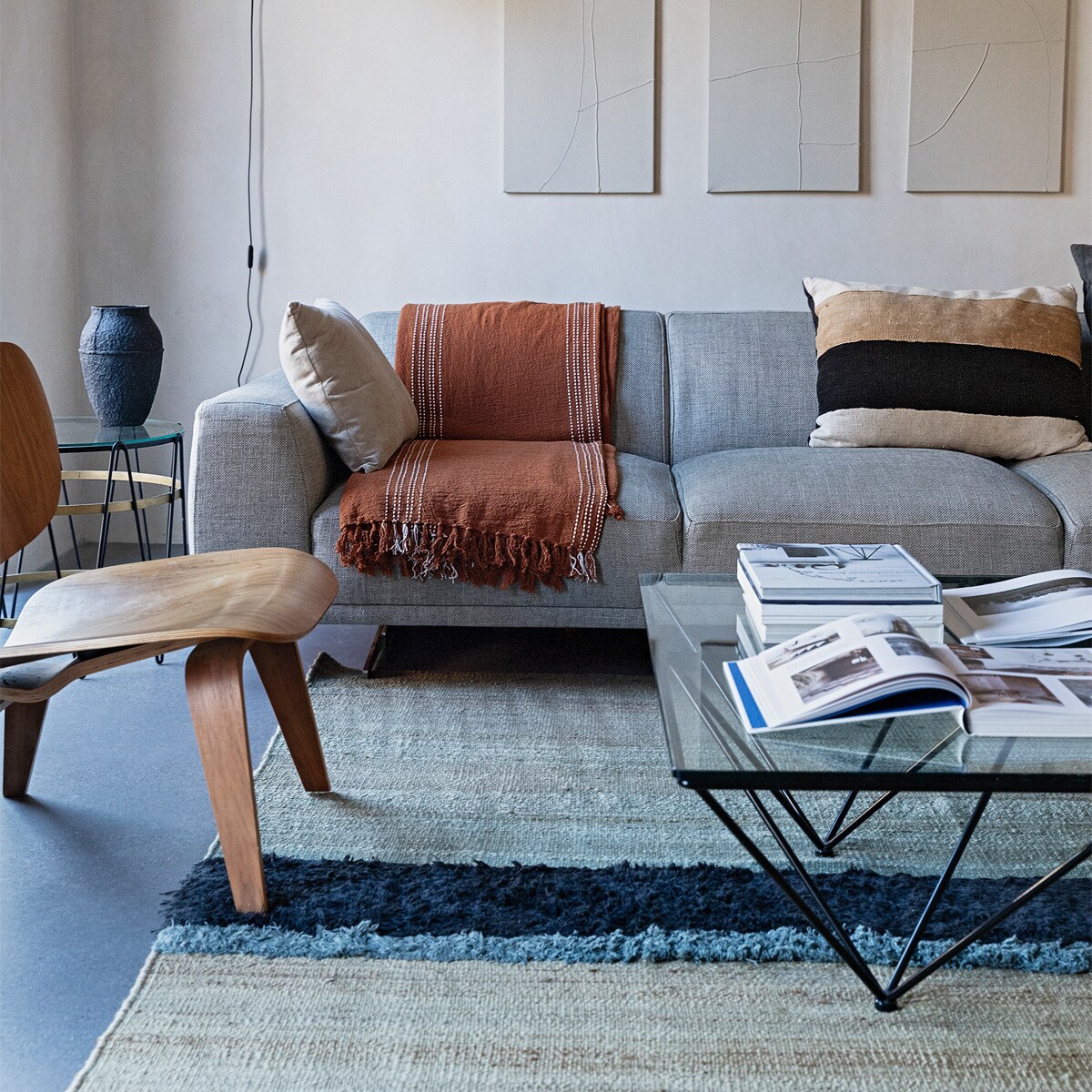 Kalkverf Mykonos van Bauwerk Colour, kunstwerken Vivienne Uzozie, wandlamp mini Lampe de Marseille van Nemo. Eames-stoel LCW Lounge Chair van Vitra. Bijzettafel Paul Lelieveld, vaas Serax. Bank Louter Design, beige kussen Zara Home, plaid H&amp;M Home, gestreept kussen The Corner Studio. Salontafel vintage Alanda Paolo Piva.