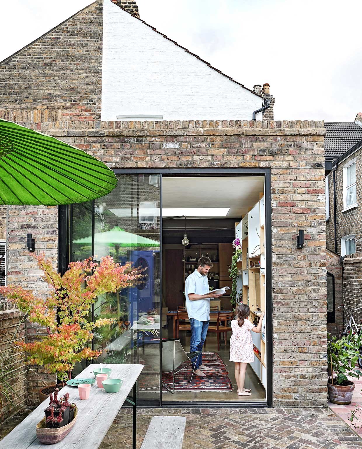 Grote schuifdeuren geven toegang tot de tuin en zorgen voor veel daglicht in dit kleine huis. Sean staat voor een van zijn op maat gemaakte kasten, samen met dochter Freya. Parasol, tafel en bank Scp. NEE | vtwonen binnenkijken special 03-2020 | styling: someday studio | fotografie: Elsa Young/bureaux