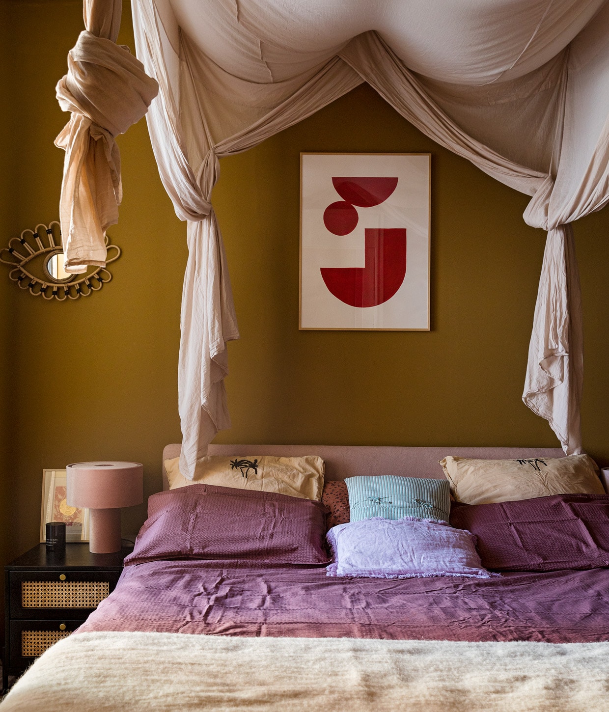 Muurverf Nova van Praxis, spiegel oog en lila kussen Madam Stoltz, kunstwerk Hôtel Magique, bed Auping, nachtkastje en tafellamp Westwing, kunstwerk op nachtkast Amber de Vreng, paars dekbedovertrek en oranje kussenslopen Crisp Sheets, blauwe kussens Khasto, plaid The Souks, klamboe Numero 74.