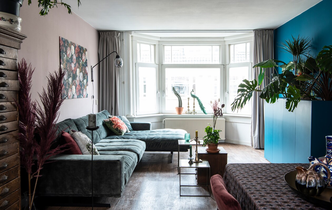 Bank, bijzettafels, cactus, plant op ladekast, kaarsenstandaards op tafel, schaal en veren Blooming Interior. Kussens Vanilla Fly, lamp BePureHome. Pauw By-Boo. Ladekast gemaakt door de opa van een vriend. Herkomst gordijnen onbekend. Kaarsenstandaards voor het raam IJ-Hallen. Het zeil lag er al. Muurverf Light Peachblossom 3 van Little Greene. Kunstwerk zelfgemaakt. Gordijnen Hema.