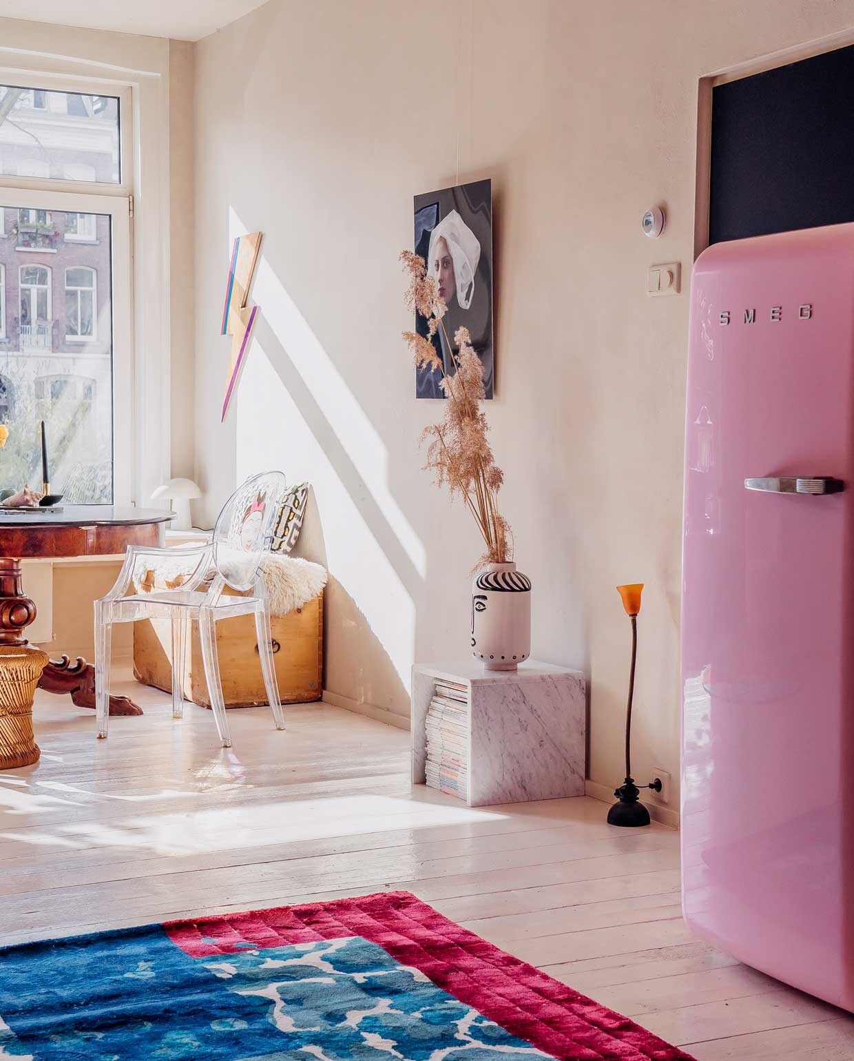 Vloerkleed Sudio Vaycay, koelkast Smeg, tafel, kist en vaas, erfstukken. Krukje Marktplaats. Transparante stoel Louis Ghost van Kartell. Foto aan de wand uit de serie ‘Paula’ van Hendrik Kerstens. Bliksemschicht via Bride &amp; Wolfe. Vachtje gekocht op Noordermarkt. Kussen Living Stories.