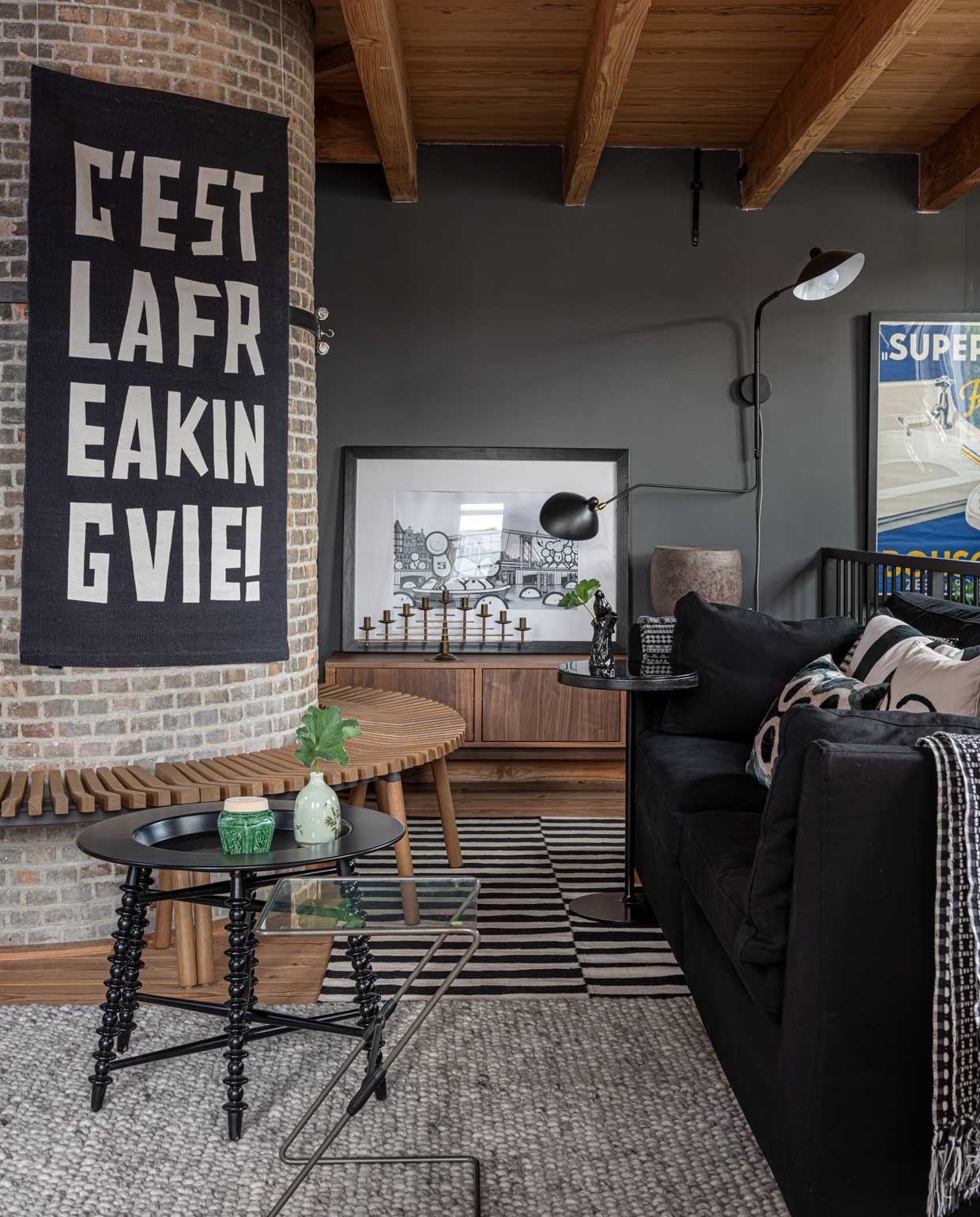 De zithoek boven. Kussen Winky van Urban Outfitters, kussen ervoor Piece of Pillow, kussen met planten­motief cadeau gekregen. Glazen bijzettafel vintage. Groene vaas en gemberpot kringloopwinkel. Kandelaar Dan Present ltd, litho The London Police, keramieken object gemaakt door Marcels moeder, kunstenares Riek Sanderse. Vintage Bousquet reclameposter Marktplaats. Witte pot met olifant & klevering. Muurlamp Serge Mouille. Kleed met tekst op schoorsteen H&M Home. Muurverf Industrial Grey van Flexa. Overige Ikea. vtwonen binnenkijkspecial 12-2020 | styling Sharon Roest | fotografie Caroline Martinot
