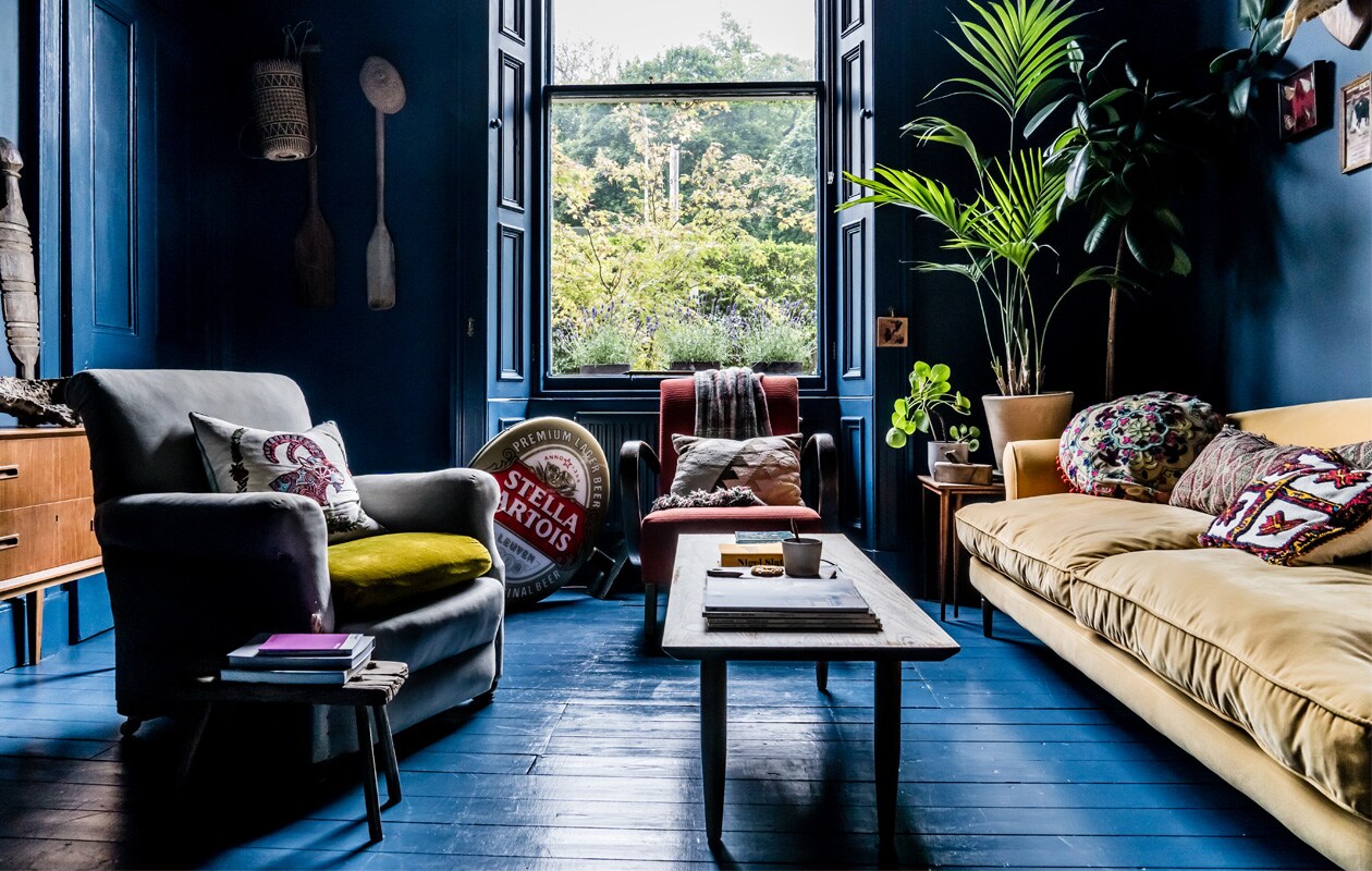 Verf op vloer, wanden en plafond Hague Blue van Farrow &amp; Ball. Rode fauteuil van Jindrich Halabala uit de jaren 30, vintage salontafel uit de jaren 60, peddels uit Inonesië, kussens meegebracht uit verschillende landen. Bijzettafels en Stella Artois-lichtreclame vintage, oude grijze fauteuil uit een winkel in Portobello. De fluwelen driezitsbank is een van de weinige nieuwe stukken. Vergelijkbare banken te koop bij o.a. Karwei, Flinders en Westwing Now.
