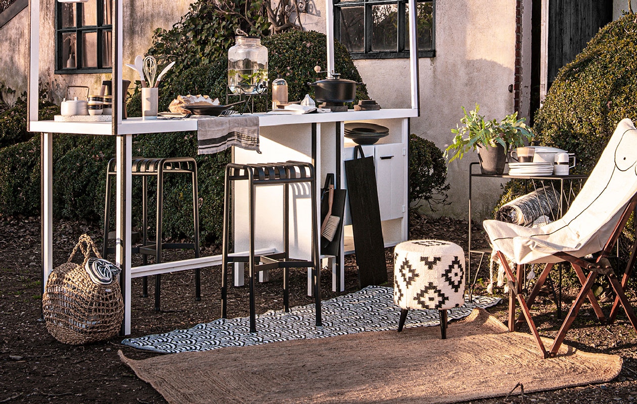 Buitenkeuken van Combitex op maat gemaakt (com-anders.nl). Zwart-wit buitenkleed van Mica Decorations, zwarte trolley Use van House Doctor, 64x40x81 cm, broodplank van HKliving, 15×55 cm, 25×45 cm, zwarte borden van HKliving, mokken Kyoto wit met zwart van HKliving, wit bord Bold &amp; Basic met palmprint van HKliving, Ø 26,5 cm, bruin-zwarte cappuccino mok met oor van HKliving, 0,3 l, zwarte braad/stoofpan Signature van Le Creuset, Ø 24 cm, waterdispenser met zwart onderstel van Madam Stoltz, 18 l (vtwonen.nl). Witte braadpan Senior met deksel, 5 l, witte kandelaar aan zijkant keuken, hoog 28 cm, stof boven keuken, 150 cm breed (ikea.nl). Beige kaars, poef met print, langwerpig beige kleed, kleed opgerold in trolley, zwarte plantenpot, gevlochten tas Ibiza, kleed in tas, zwarte stalen barkruk geschikt voor buiten (loods5.nl). Fles met zeepsop, receptenboek, uivormig potje, koffiemaker zwart, spatel en keukenlepels, stoffer-en-blik, bus poedersuiker met handvat op trolley (zarahome.com). Stoel met getekend lichaam (anna-nina.nl). Ronde slinger met streep (sukha.nl). Bruine theedoek met zwarte streep (maisonnl.com). Bruine pot waar lepels in zitten (annemiekebootsceramics.nl). Witte theepot van HKliving (deens.nl). Terrazzo serveerschaal van HKliving (fonq.nl).