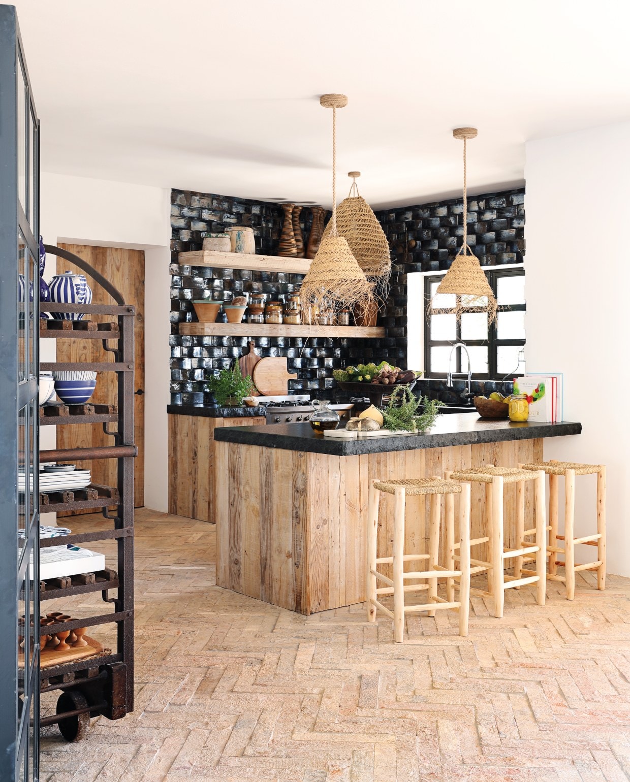 De keuken kreeg diepdonkerblauwe keramische tegels met een zweem zwart erin. De luxe glans steekt mooi af bij de natuurlijke materialen. Hanglampen Hubert Zandberg Interiors, barkrukken Mona Market. De aanrechtbladen zijn van zwart leisteen. vtwonen 08-2020 | binnenkijken buitenland ibiza | Fotografie Elsa Young