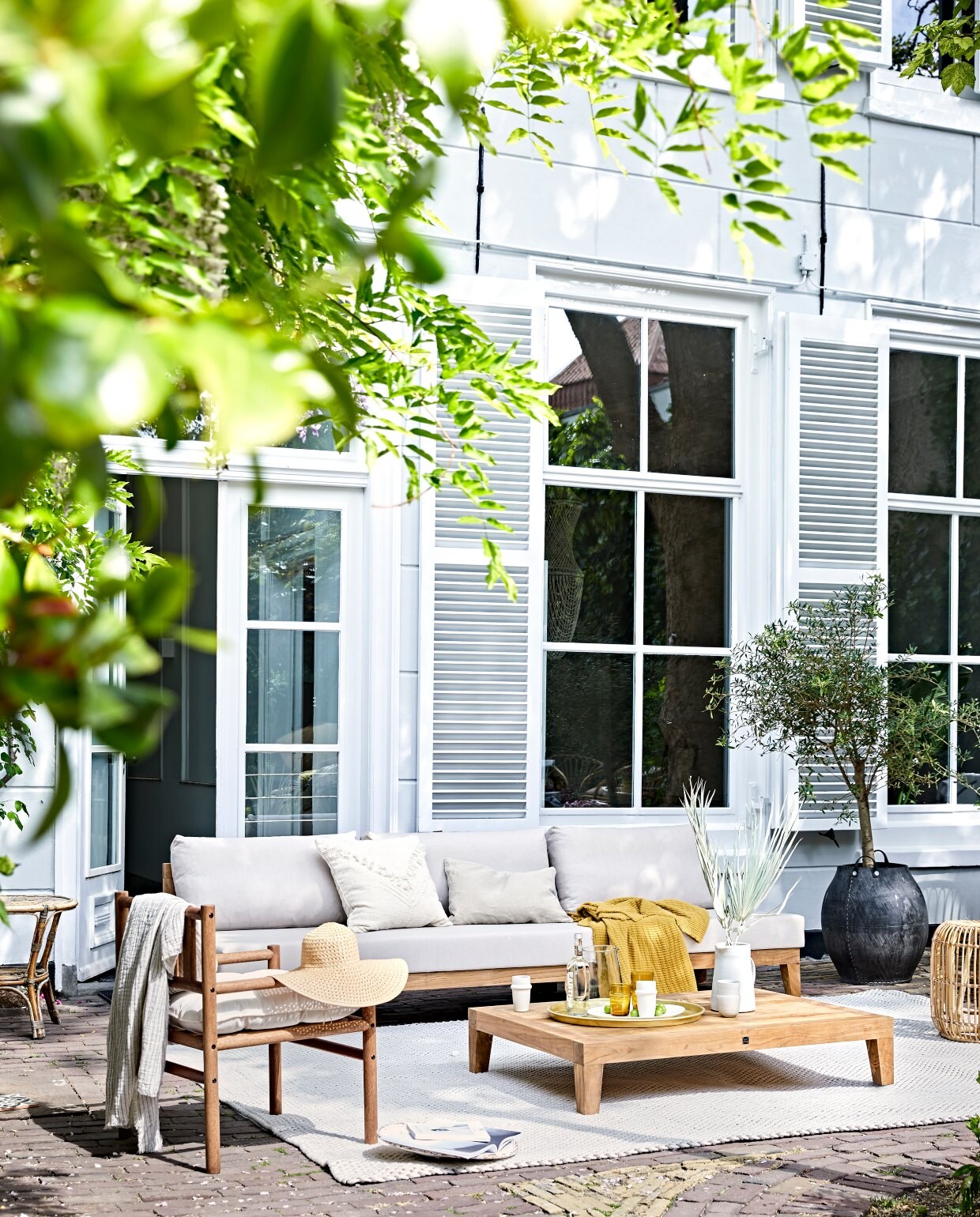 Buitenbank Koen bestaande uit een 2-zitselement met kussens,180×90 cm € 1795,-, en 1-zitselement met kussens, 90×90 cm € 995,-, buitenvloerkleed 250×350 cm € 1395,-, tafel 90×90 cm € 595,- (royaldesign.nl). Marantiehouten fauteuil met kussens € 149,- (hm.com). Okergele plaid € 82,75, zandkleurige plaid € 103,50, Marokkaanse bekers € 5,50, wit kussen met franjes, 50×50 cm € 55,25 (madamstoltz.dk). Gouden dienblad € 49,50, glazen kan € 33,50 (weldaad.com). Keramieken kan € 99,- (sukha.nl). Zandkleurig linnen kussen € 34,95 (iblaursen.nl). Wit bord € 36,50, okergele waterglazen € 7,99 (nordal.eu). Overig privébezit. vtwonen 07-2020 | Styling Cleo Scheulderman | Fotografie Jeroen van der Spek