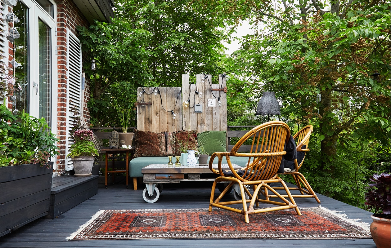 Het terras heeft verschillende niveaus en verbindt het huis met de tuin. Op het bovendek een oud loungebed bekleed met waterafstotende stof, een vintage rieten stoel en een zelfgemaakte pallet tafel op wielen met tegels van Arttiles. Erachter een paar oude deuren die een nieuw leven kregen als schutting. De lamp is van Ikea, de lichtketting kocht Trine bij de lokale bouwmarkt. Het grote tapijt vonden ze tijdens een reis naar Iran. De kussens zijn zelfgemaakt. Kleine lantaarn uit Marokko, tafeltje vintage. In de zelfgebouwde plantenbak staan lupines. Tegen de luiken klimt een clematis. vtwonen tuin special 1 | hoog leef terras met loungebank en rotan stoelen