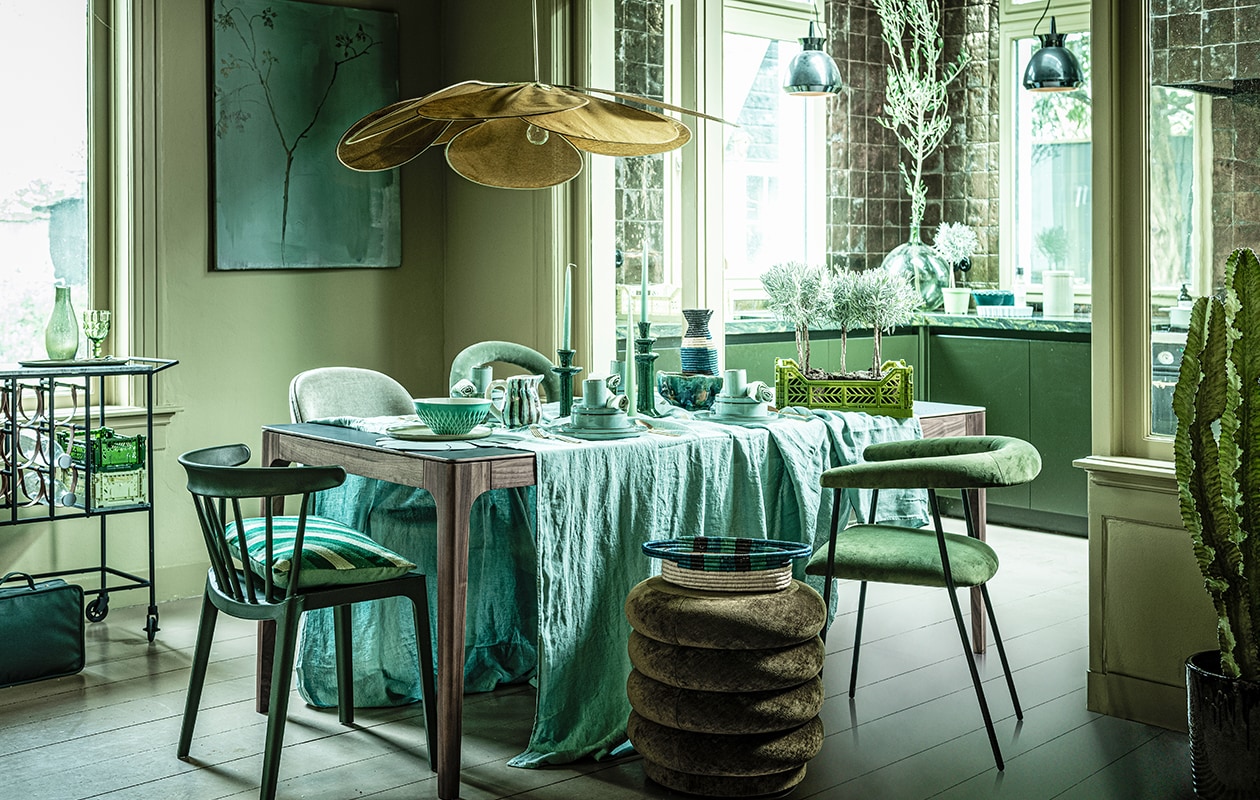 Tafel Rikke walnoot met Fenix blad, 160x90x76 cm (demachinekamer.nl). Poef (fermliving.com). Velours stoel links Gubi Beetle, glazen fles op trolley (homestede.nl). Velours stoel rechtsachter (covanderhorst.nl). Houten stoel Bliss van Woood, velours stoel rechtsvoor Haily van Dutchbone, trolley Mil van Dutchbone, rond bord Surface van Serax ovalen tapasbord Surface van Serax, braadpan Surface van Serax, waterglazen Raami van Iitala, goudkleurig bestek Amefa vintage, taartvorm tarte tatin van Jamie Oliver (vtwonen.nl). Wijnglas op trolley (zarahome.com). Waterflessen, krat klein, krat groot, kan gestreept, kandelaars en kaarsen, opgestapeld: kopjes klein, borden, schaaltjes middel, groenblauwe gevlekte schaal, voorraadpot, groene schaal op keukenblad (loods5.nl). Linnen tafellaken, 320×270 cm, mand, hoge kan (couleurlocale.eu). Koffertje, lamp, Ø 120 cm (archive-store.nl). Kussen (hinck.nl). Linnen servetten (hm.com). Grote vaas (oudebouwmaterialen.nl). Groene schaal op tafel (ottomania.nl). Placemat (bijenkorf.nl). Plantenpot (uncwardrobe.com). Schilderij Branch on a green canvas, 80×100 cm (maartjevandennoort.nl).