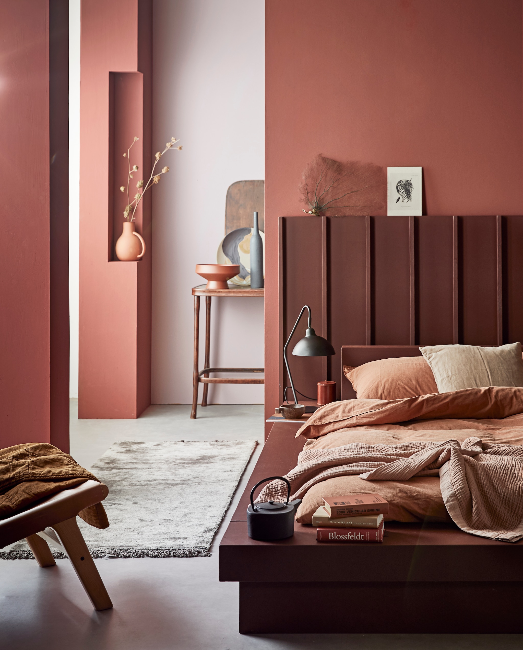 Muurverf Red Earth (voorste wanden en kolom), Washed Lilac (achterwand), lakverf Terra (bed en headboard)M.m.v com-anders.nl (op maat gemaakt headboard en bed), vtwonen.nl (tafellamp Orion, beddengoed Natural Stone in kleur Terra, roze sprei en stoel van hout en leer uit de vtwonen collectie, groot bord met gezicht van Ferm Living), nordal.eu (terracotta vaas met oor, vloerkleed, grote terracotta schaal, theepot, karpet), iblaursen.com (quilt op stoel, roze linnen kussen), maisonnl.com (bruin rond leren potje, bruin boek). Overig privébezit.