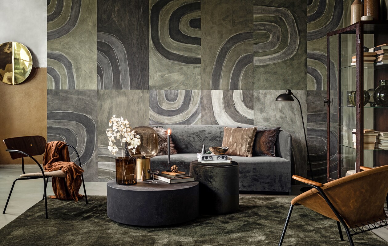 Bank Tearoom Sofa van Menu, 190x78x70 cm (lxhxd) (designtales.nl). Behang op wand mural Agata (wallanddeco.com). Messing wandobject, fauteuil met suède zitting en houten armsteunen (goeds.nl). Stoel, bruin kussen met reliëf (homestede.nl). Bruine plaid (hm.com). Lage ronde bijzettafel betonlook van Woood, Ø 86 cm, groene velours poef, Ø 46 cm, bruine glazen vaas van Madam Stoltz, tafellamp met grijs glas en messing poot van House Doctor, zwarte metalen kandelaar van Eno Studio, lamswollen sierkussen van Bloomingville (vtwonen.nl). Marmeren bomen, boek met ogen, donkergroen notitieboek, okergeel notitieboek (klevering.nl). Schaal van hoorn (letoile.store). Beige kussen met vleug, groen vloerkleed (covanderhorst.nl). Zwarte vloerlamp (coosdewitwonen.nl). Vitrinekast (vandijkenko.nl). Op en in de kast van boven naar beneden: beige stenen flesvaas van Madam Stoltz, bruine stenen flesvaas van Madam Stoltz (vtwonen.nl). Marmeren potje met deksel, grijs geribbelde glazen vaas, grijze glazen bolvaas (hm.com). Zwart mini-vaasje, groene stenen vaas (fleurmonde.com). Overig privébezit.