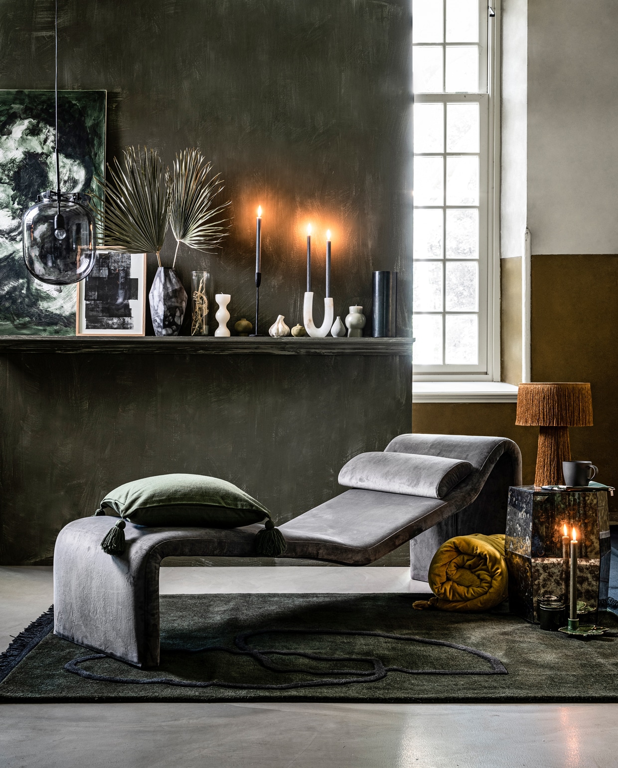Grijs fluwelen daybed (polspotten.nl). Verf op wand Fresco kalkverf in kleur Belgian Wilderness (pure-original.nl). Op de plank: schilderij door kunstschilder Jan Wiedijk (danielleverheul.com). Poster houten lijst, zwartmetalen kandelaar (thingsilikethingsilove.com). Gemarmerde glazen vaas, cilindervaas met droogbloemen, wit marmeren kaarsenhouder (loods5.nl). Blokkaarsen (klevering.nl). Mini-vaasjes in groen, wit en grijs (fleurmonde.com). Antraciet cilinderlamp van Serax (vtwonen.nl). Op de voorgrond: grijze glazen hanglamp van House Doctor mosterdgele tafellamp met kwastjes van Madam Stoltz, groen vloerkleed met blauw lijnenspel van Ferm Living, 200×140 cm (lxb) (vtwonen.nl). Opgerold geel fluwelen zitkussen gespiegelde bijzettafel (loods5.nl). Etui wit met groene stippen (klevering.nl). Groen kussen met kwasten, grijze mok , bladvormige kandelaar (hm.com). Groene stenen pot met deksel (thingsilikethingsilove.com). Overig privébezit.