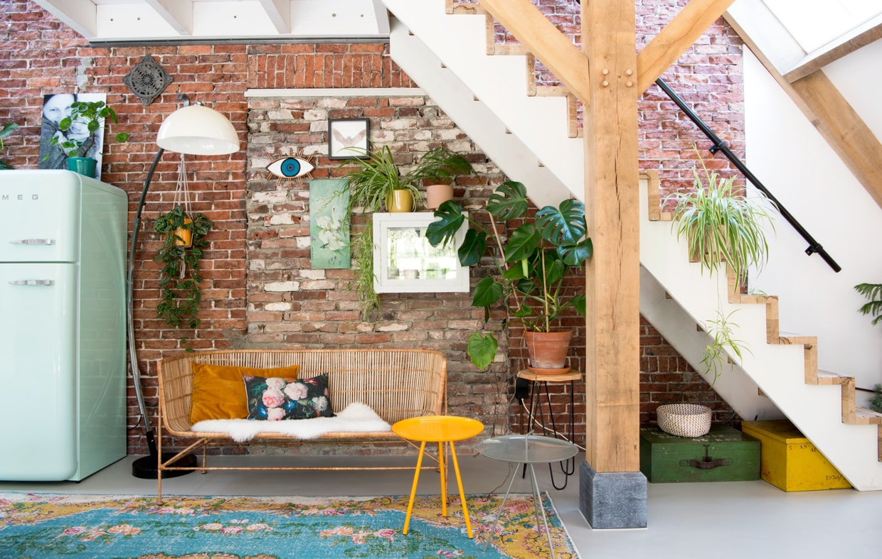 Booglamp van een kringloopwinkel. Bankje van House Doctor via vtwonen.nl, geel tafeltje van Zuiver, beker erop van Bloomingville, kastje aan de muur tweedehands, oker kussen van Limelight, kussen met rozen van Loods 5, oog aan de muur van Umasqu, vleermuis van vlindersinlijst.nl, zwanenschilderij van de kofferbaksale die ‘s zomers elke zondag wordt gehouden in Schoorl, bloempotten van Dille &amp; Kamille.