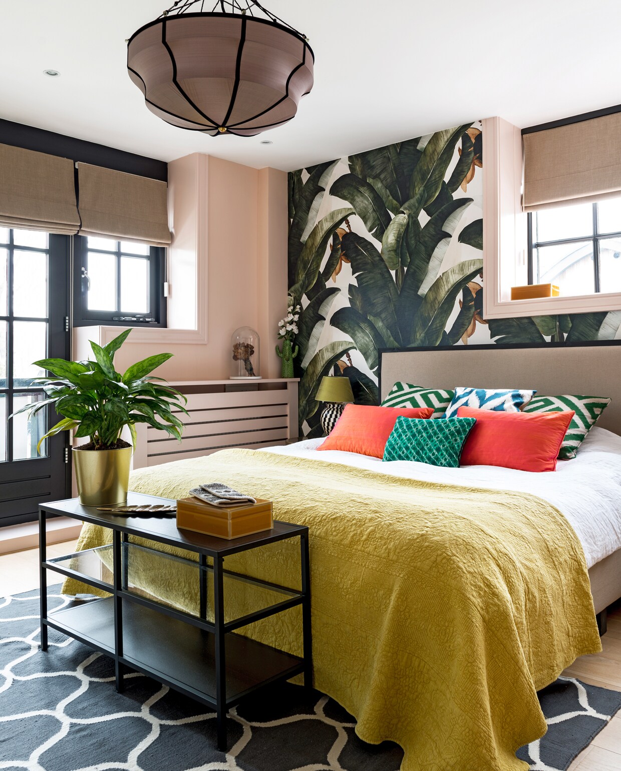 Behang van Lemon. Hanglamp van Gong. Headboard bed van Studio Sophie. Sprei van Zara Home. Groen kussen van Elitis, rode kussens van de groothandel en Ikat kussens van Eveline Interieur. Zwarte sidetable, vloerkleed en plantenpot Ikea. Glazen stolp met gedroogd bruidsboeket. Lamp met zwart-witte voet van Flamant. Roze verf Pink Ground van Farrow &amp; Ball. vtwonen special binnenkijken 12-2019 | styling Lindividu | fotografie Stan Koolen, CocoFeatures