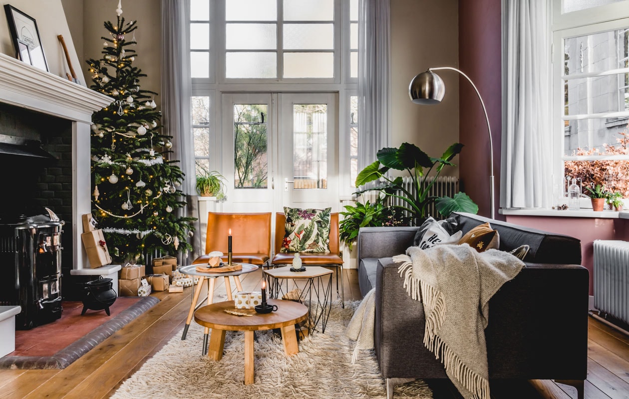 De grote boom staat mooi naast de bakstenen schouw, die met plamuur en verf werd opgefrist. Elk jaar komen er een paar nieuwe versierinkjes bij voor in de boom. Bijzettafels van Xenos en Zuiver, bruine fauteuils van Kwantum, vloerkleed van Ikea. vtwonen 12-2019 | productie Features &amp; More | styling Wilma Custers | fotografie Ivar Janssen