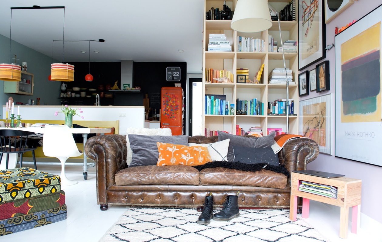 Rond het trapgat is een kast gebouwd voor keukenspullen en boeken. Jet en Michiel lieten hem maken bij dewinkeldiewerkt.nl. De vloer is gemaakt van duurzame eikenhouten duoplanken, gekocht bij Fairwood. Kunst van boven naar beneden: Tate Gallery By Tube poster gekocht via The London Transport Museum, foto Frida Kahlo, kunstwerkje gemaakt door de opa van Michiel, onder een eigen werk van Jet. Grijze kussens bank Madam Stoltz, oranje kussen The Pillow Room. Zwarte eetkamerstoel Hay. vtwonen 12 binnenkijken special 2019 | Styling Jet van der Graaf | Fotografie Trui Heinhuis | Tekst Caroline Westdijk