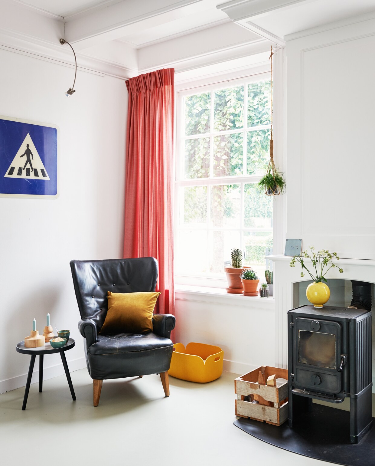 Stoel Artifort. Kussen Particolare, houten kandelaars Marimekko, gele opbergdoos Restore door Muuto, gele vaas Kähler, alles via Nio &amp; Elva. Bijzettafeltje Karwei. Houtkachel Mørse. Tegeltje op schoorsteenmantel Storytiles. Vintage verkeersbord via Living Once. Gordijnen via Post Woonadvies. Plafondlampje hing al in het huis. plantenhanger via De Haas Bloemen.