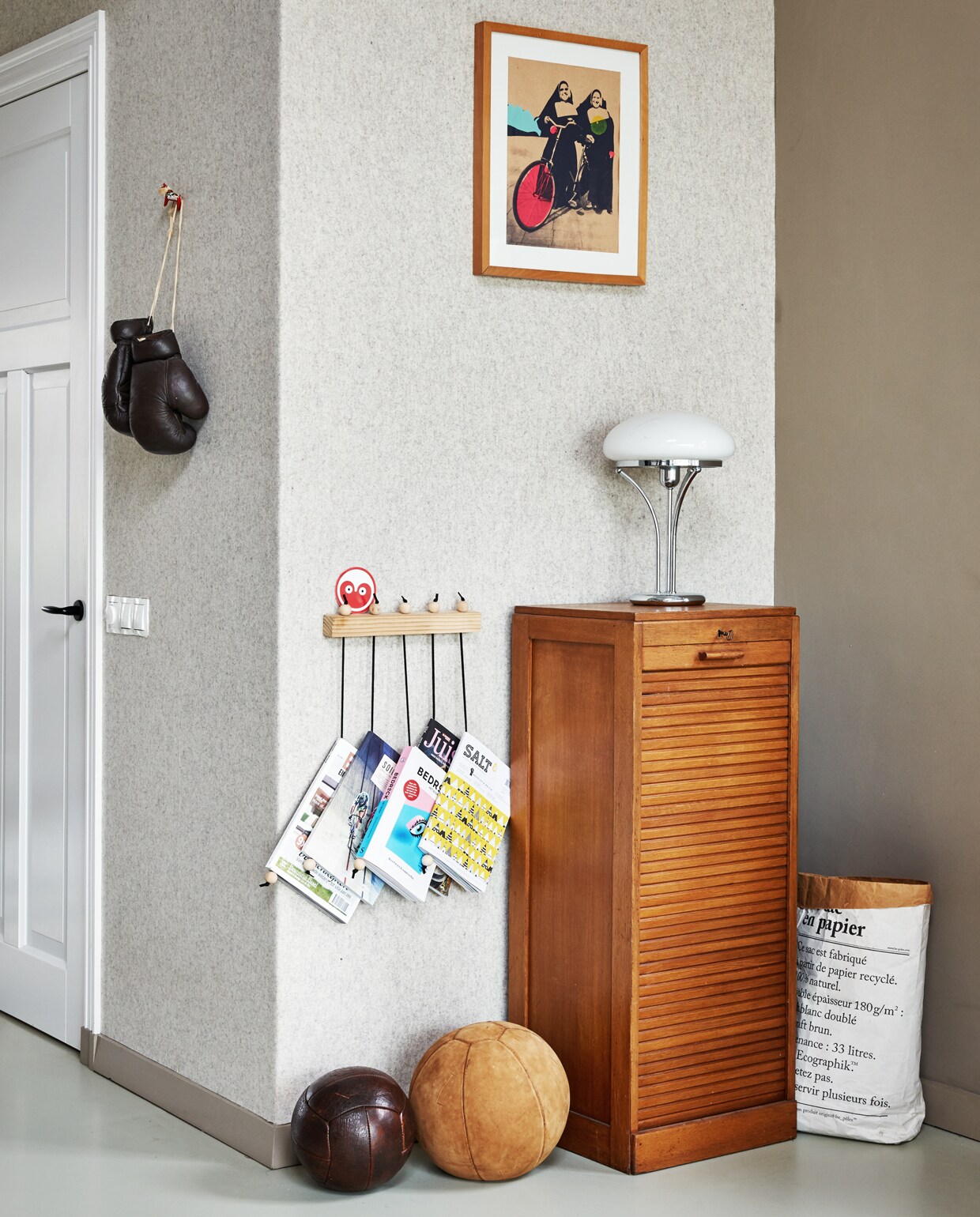 Op de muur lchtgrijs gemêleerd vilt kleur naturel/32 via wolvilt.nl. Tijdschriftenrek en tafellamp Hema, sticker Leeuwarden European Cultural Capital 2018. Vintage ballen via Living Once. Zeefdruk door Saskia. Papieren mand Le sac en papier. Bokshandschoenen cadeau van ex-boxer. Vintage kastje is gekocht bij Living Once.
