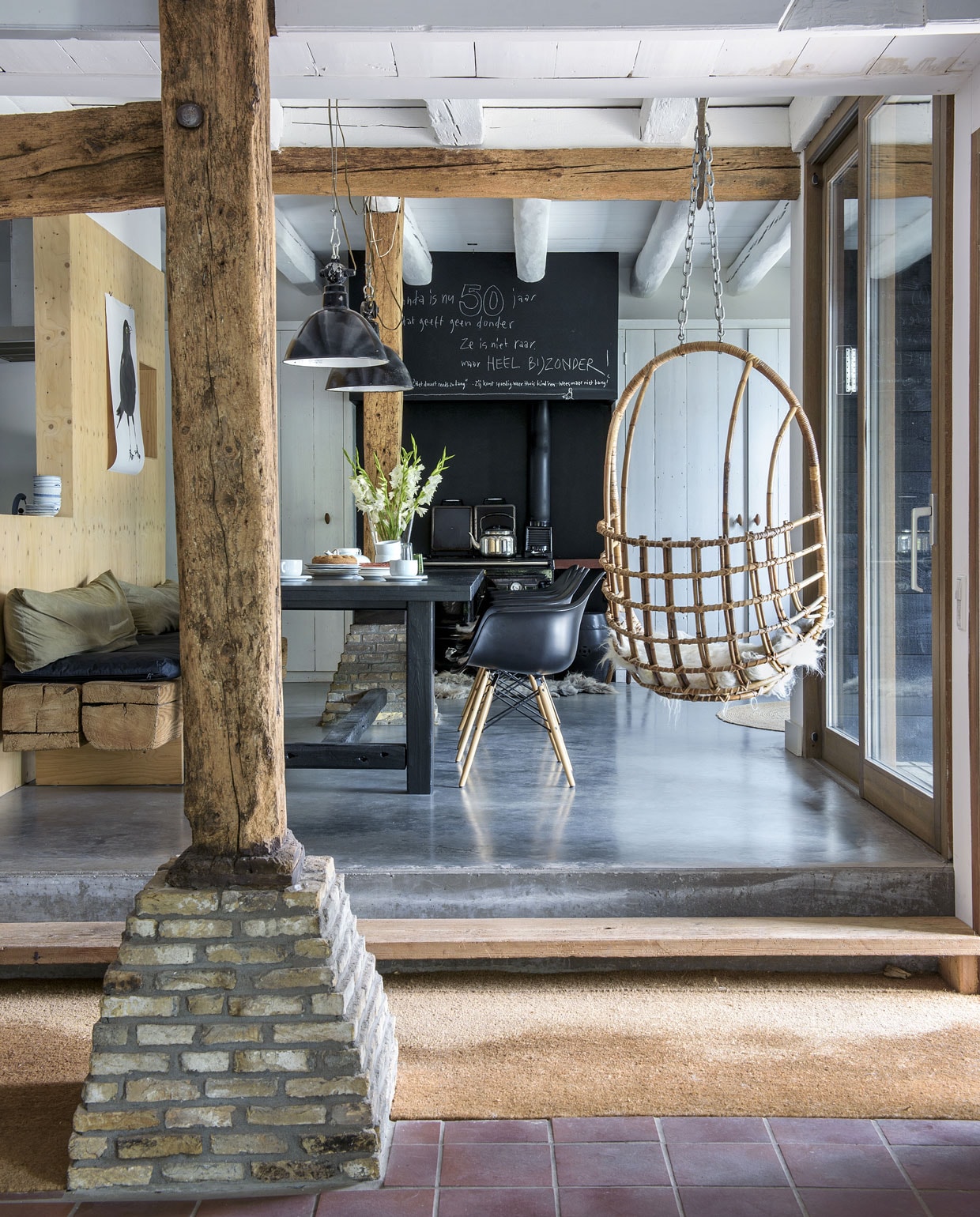 De tafel en het bankje werden zelfgemaakt van materialen uit het huis. De zwarte stoelen zijn van Eames. Lampen van Buitengewoon Brocante. Illustratie Maartje van den Noort. De oude staander loopt door tot aan het dak. De vloer is van beton. Vanuit de keuken loop je zó door de schuifpui naar de patio, waar het goed chillen is in de hangstoel van Sissy Boy Homeland. vtwonen 11-2019 | styling Monique van der Pauw | fotografie Ton Bouwer