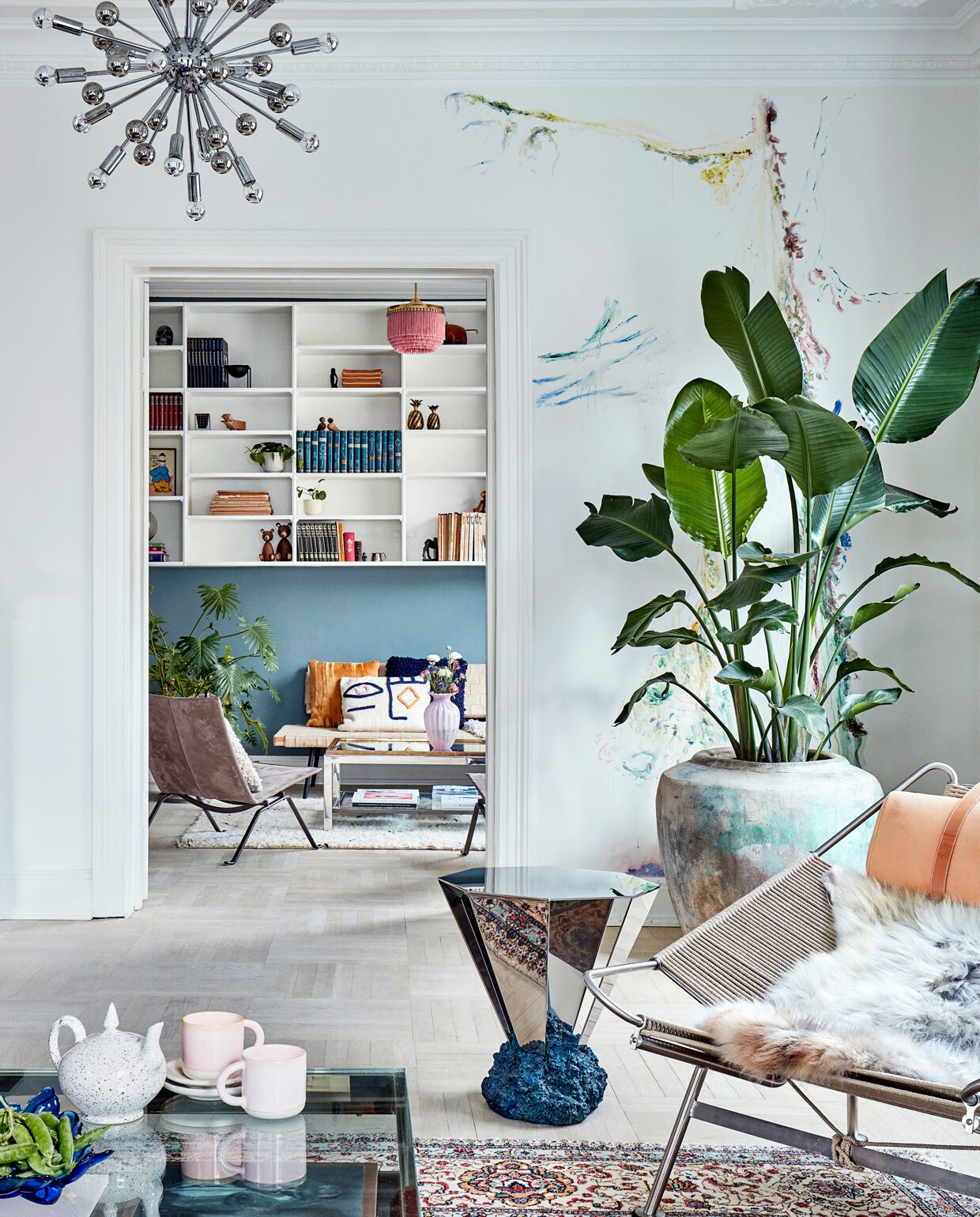 Kijkje vanuit de zithoek richting de bibliotheek met zijn originele open wandkast. Op de voorgrond de Flagline Chair van Hans Wegner, de Diamant Table van Fos, lamp Sputnik van Paul de Haan en een vintage salontafel van Belgo Chrom. Op de achtergrond stoel PK 22 van Poul Kjaerholm, een vintage salontafel van Romeo Rega en een daybed van Ikea. De wand rechts is beschilderd door kunstenaar Jacob Steen.