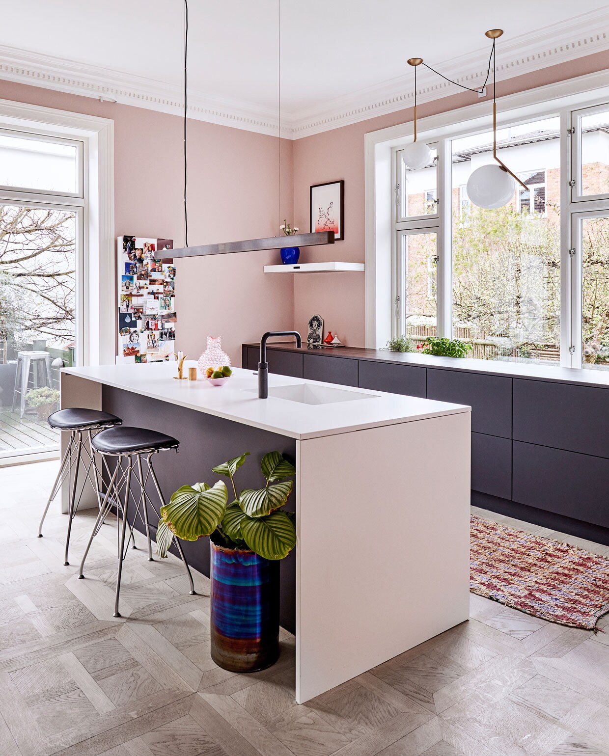 Bleekroze wanden verrassen in de keuken, die wordt gedomineerd door strakke units gemaakt door het Deense bedrijf UnoForm. Het blad is van composiet. In werklicht wordt voorzien door een handgemaakte donker koperen lamp van Anour, de bollampen IC – met koperen armatuur – zijn van Flos. De krukken zijn van Overgaard &amp; Dyrman, de geglazuurde vaas is een ontwerp van Tom Dixon voor Roomstore. Op de vloer ligt een Boucherouite, een traditioneel, handgeknoopt kleed uit Marokko.