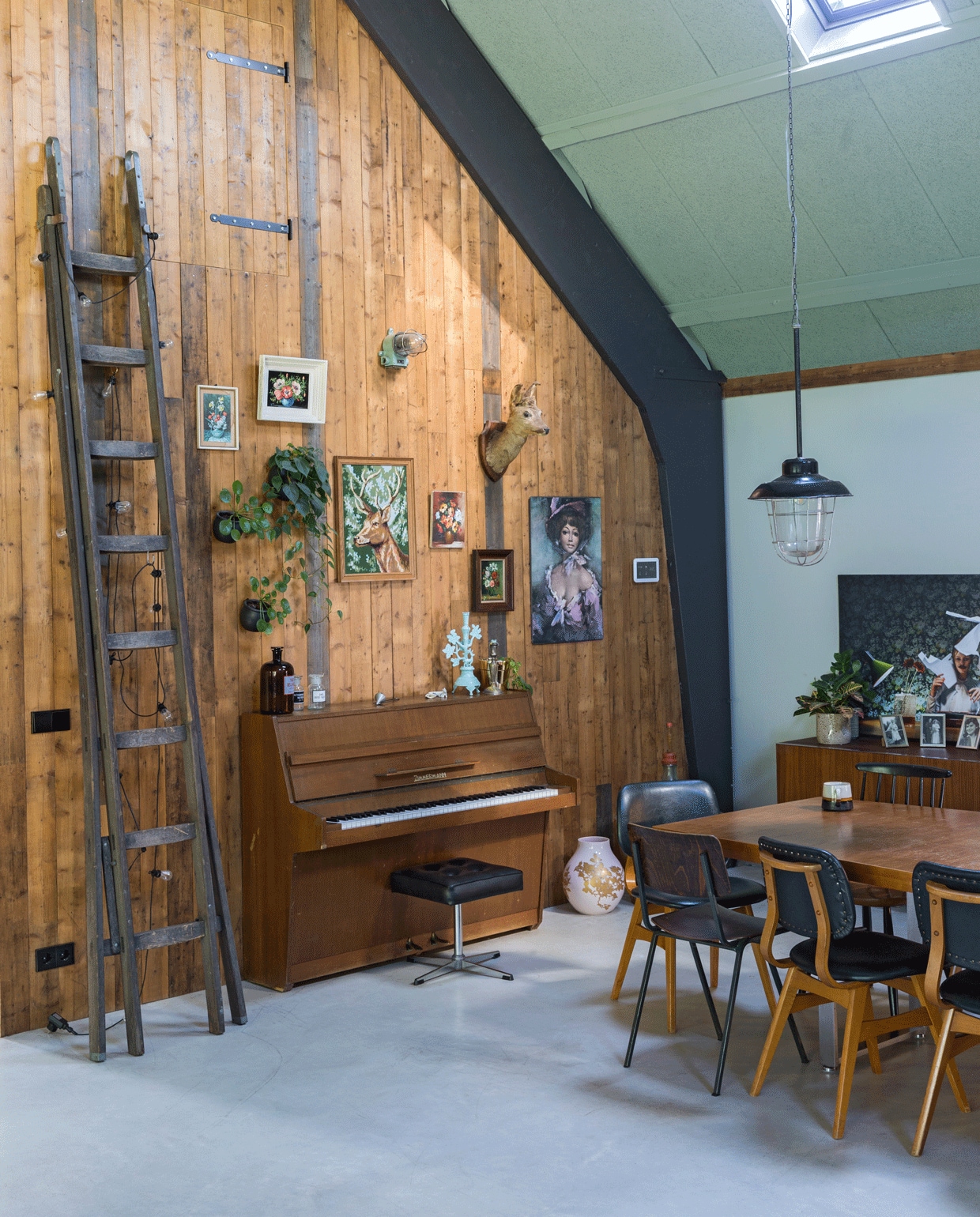 De houten wanden zijn gemaakt van een partij verweerd dakbeschot. De ladder tikte Wende op de kop via Facebook, het lichtsnoer eraan is van Intratuin. Ljstjes aan de wand via brocantemarkten in Frankrijk. De piano is van een collega geweest, erop flessen van Davidowski, kandelaar Rice. Tafel van Mooi Muf, eromheen leren stoelen van Montmartre, een brocantemarkt in Gouda, stalen stoel Davidowski, spijlenstoel Tony’s Garage Sale. Grote foto op het dressoir van Hugo. Hanglamp Vroeger &amp; Toen.