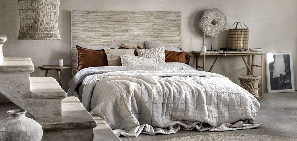 Wand: behang Reunited 372556 hoofdeinde: 140×180 cm (hxb), behang Natural Wallcoverings II 389517 (eijffinger.com). Linnen dekbedovertrek 240×220 cm, roestbruine linnen kussenhoezen 50×90 cm (hm.com). Kussen 60×60 cm, kussen 50×50 cm (mlfabrics.nl). Macramé kussen (woondock.nl). Rechthoekig kussen met leerkoord, zandstenen kruik (stylexclusief.nl). Naturel linnen sprei prijs op aanvraag, schaal rechts Broste Copenhagen, poster (loftloft.nl). Wissellijst (ikea.nl). Bijzettafel links Tripod (hetkabinet.nl). Vouwtafel van bamboe, rieten taboeret (woondock.nl). Bamboe lantaarn (stylexclusief.nl). Schelpencirkel op standaard, visfuik-lamp links (manggisliving.nl). Vloerkleed Ø 200 cm (kwantum.nl). Mok privébezit.
