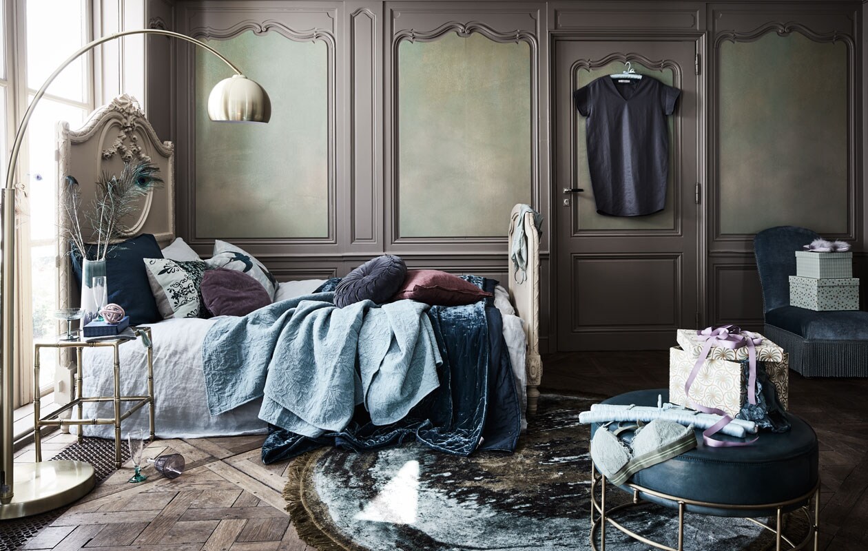 Bed 205x110x114 cm (lxbxh), glas met groene voet (wildschut-antiques.com). Staande lamp goud, vaas gekleurd op nachtkast, paarse kunstvorm op nachtkastje, blauw rond kussen, kussen donker/lichtgroen met beige achterkant, dekbedovertrek linnen 220×240 cm, rond vloerkleed Bold Monkey Ø 175 cm, etui (loods5.nl). Poef blauw (www.purefurniture.com). Kledinghanger, sloffen, paars glas, plaid reliëf (zarahome.com). Kussen rond paars, kussen vierkant paars, lint paars (bungalow.dk). Stoel rechts in de hoek (pietjonker.nl). Drinkglas groen, tafeltje goud, fluwelen plaid op bed, groene kussenhoes hoort bij dekbed tweepersoons (hm.com). Kussen met vogel van John Derian, behang Suisai 140×300 cm (wva.nl). Pauwenveren (harlequin-lisse.biz). Jurk aan kast (sukha.nl). Beha Love Stories, onderbroek Love Stories, body over bed (maisonnl.com).