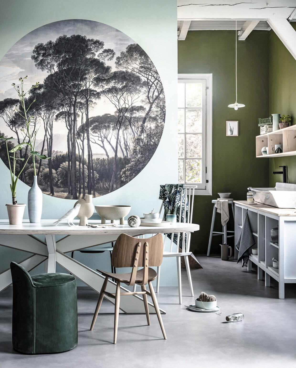 Vloer van Novilon vtwonen in de kleur Kiezel (vtwonen.nl). Cirkelvormig behang Golden Age Landscape van Kek Amsterdam, Ø 190 cm, verf op de achterwand Olive Colour 72 van Little Greene, rond het behang Aquamarine-mid 284 van Little Greene (deruverfenbehang.nl). Ovale tafel van Arp (homestock.nl). Op tafel: kamerplanten (klavertjevijf.com). Lichtblauwe vaas (zarahome.com). Lamp Bird (homestock.nl). Beige bolvaas, witte schaal op drie poten (artilleriet.se). Kommen en schalen van, witte borden, wit bestek (annemiekebootsceramics.nl). Kop met verlooptint (studioharmenelke.nl). Lichtgroene stoel achter tafel (hay-amsterdam.com). Groene kruk met rugleuning (edha-interieur.nl). Houten stoel, hoge kruk achterin (demachinekamer.nl). Witte stoel (loods5.nl). Stof erover (ikea.nl). Landelijke keuken, kraan, gootsteen en wandschap (combitex com-anders.nl). In de keuken: platte hanglamp, blauw en grijs servies (studioharm-enelke.nl). Lijst aan de muur print erin (mydeerartshop.nl). Kratten bovenop het wandschap (hay-amsterdam.com). Zeeppomp (mydeerartshop.nl). Groene theedoek (hay-amsterdam.com). Servetten van beige linnen (kniphal.nl). Marmeren vijzel (mydeerartshop.nl). Op de grond: grijze koffiemaker van Muuto (misterdesign.nl). Fles met plant (loods5.nl).