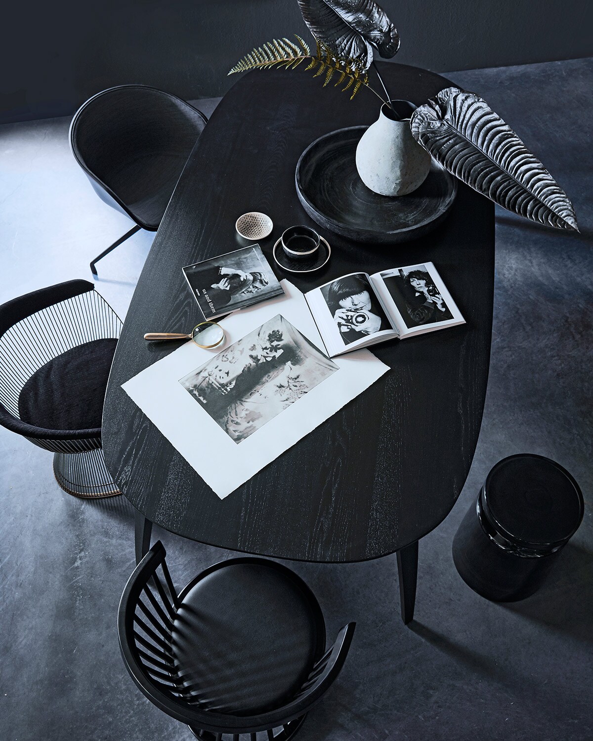 Eettafel Ellipse van Gubi, zwarte houten spijlenstoel Fan van Tom Dixon, zwarte gestoffeerde kuipstoel AAC20 van Hay (nordicnew.nl). Vintage Warren Platner stoel (demachinekamer.nl). Zwarte glazen kruk, houten schaal, vaas, schaaltje, alles van Mvn&amp;i (mvninteriors.com). Zwarte schaal en kom met witte rand (couleurlocale.eu). Fotopolymeer ets Where are you #6 van Chantal Elisabeth Ariëns, gedrukt op Velin BFK Reeves-Arches papier (chantalariens.com). Vergrootglas (zarahome.com). Boeken (taschen.com).