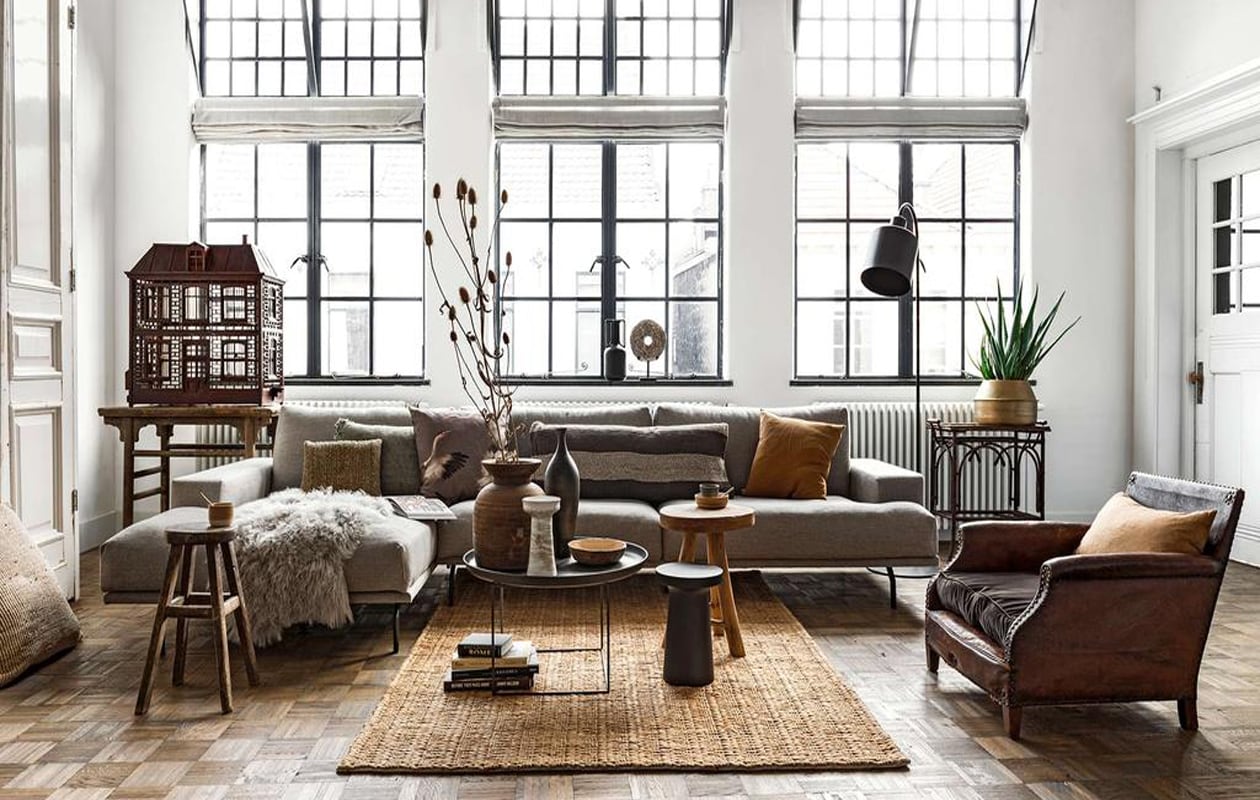 Loungebank Bobbie, ligelement, oud uniek houten tafeltje links naast de bank, zwarte metalen salontafel Norr Duke Large, hoge oude houten kruk, matzwarte vloerlamp Boris, messingkleurige vaas met daarin een aloë vera, ronde steen op voet, diverse kunstboeken, grote houten pot (loftloft.nl). Antieke vogelkooi van begin 1900, antieke leren fauteuil van begin jaren 30, antiek exotisch theetafeltje van begin jaren 30, rechts naast de bank (interieur-vivre.nl). Houten kruk Modern Earth, zwarte houten kruk Shizu, jute vloerkleed van BePureHome, Mongools schapenvacht van Lene Bjerre, kussen met vogels Aza van Claudi Design, camelkleurige kussens van By Mölle, langwerpige wengé houten carved vaas van Scapa Home, zwart kruikje van keramiek (vtwonen.nl). Zeegras kussentje Mekong Nights, grijs linnen kussen met franjes Indochine Mornings, linnen langwerpig bruin-crème gestreept kussen Mekong Nights, zeegras kussen Indochine Mornings op de vloer (aaimadewithlove.com). Cappuccinomokje Basalt Grey, mokje Walnut, lepeltjes, schaaltje Walnut (annemiekebootsceramics.nl). Bamboe schaaltje en wit vintage vaasje privébezit.