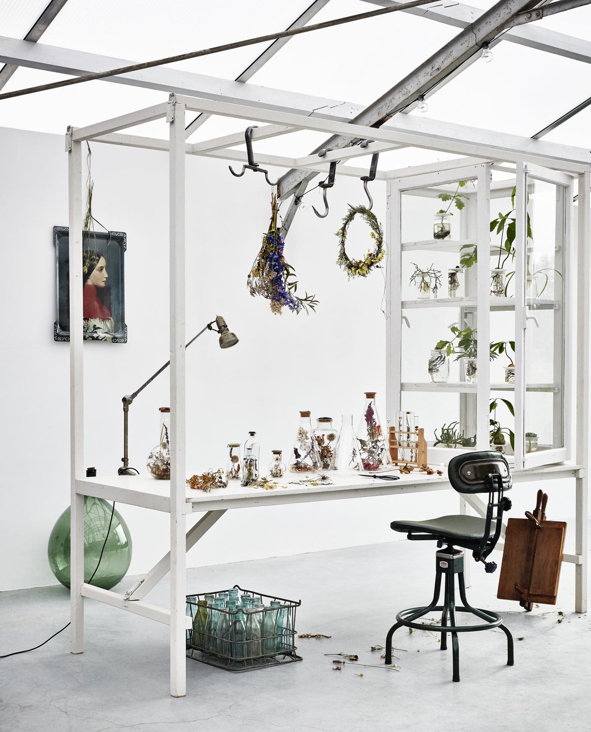 Houten werktafel met kweekkast (Combitex). Draaistoel, krat met flessen, fles, klemlamp, haken (Brût Amsterdam). Dienblad met portret van Ibride (MaisonNL). Op tafel flessen Flasks of Hope en Tubes of Hope (Field of Hope). In de glazen kast kweekschaaltjes Sprout van porselein (Sprout). Droogbloemen (Dille &amp; Kamille). Kransje en droogbloemen (Elin Wanderlüst). Bloemenpers privébezit.