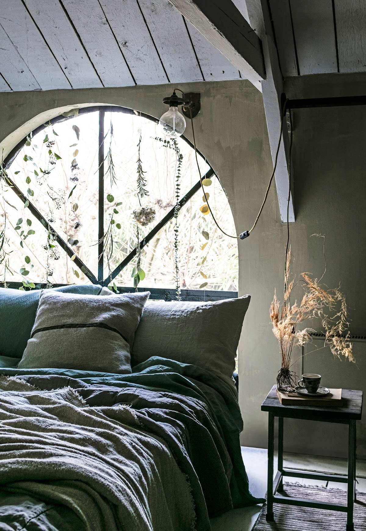 Gordijn van groen voor het raam, gedroogd takje (Tuin van Judith). Kussensloop, overige kussens op bed, linnen laken, nachtkastje, wandlamp Glowing Blossom, ontwerp van Nik Baeyens, kop-en-schotel, vloerkleed, gerafelde linnen doek op het bed (Meker Wonen).