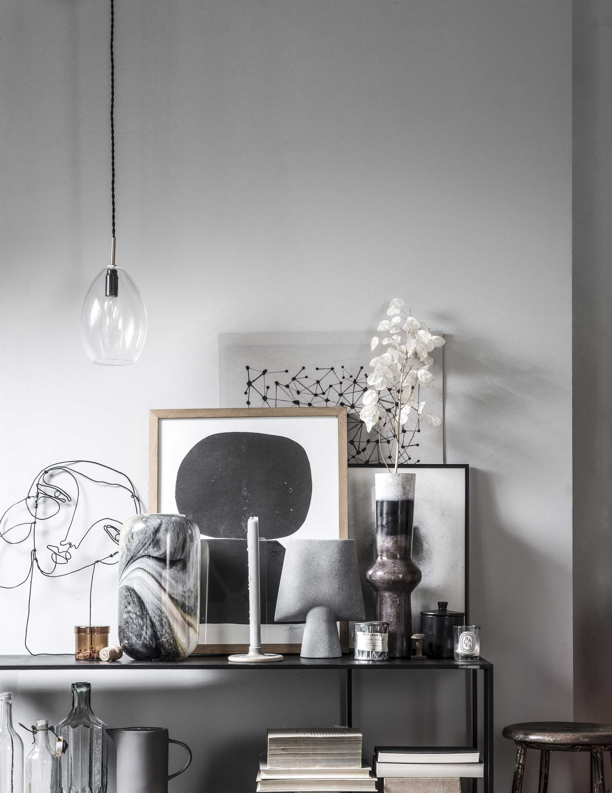 Zwarte metalen dressoirkast . Glazen hanglamp . Gezicht van metaal-draad, inclusief verzendkosten . Bruine glazen pot met deksel, grijze vaas, bruine vaas met witte bovenkant . Glazen vaas met grijze en beige waas . Kandelaar van keramiek . Le Labo geurkaars in metalen blik . Zwarte zeefdruk in houten lijst van Anne Olde Kalter . Poster Graphic Grain, zwarte aluminium lijst . Poster geprint op grijs karton . Houten kruk . Grijze glazen vaas op onderste plank . Overig privébezit.