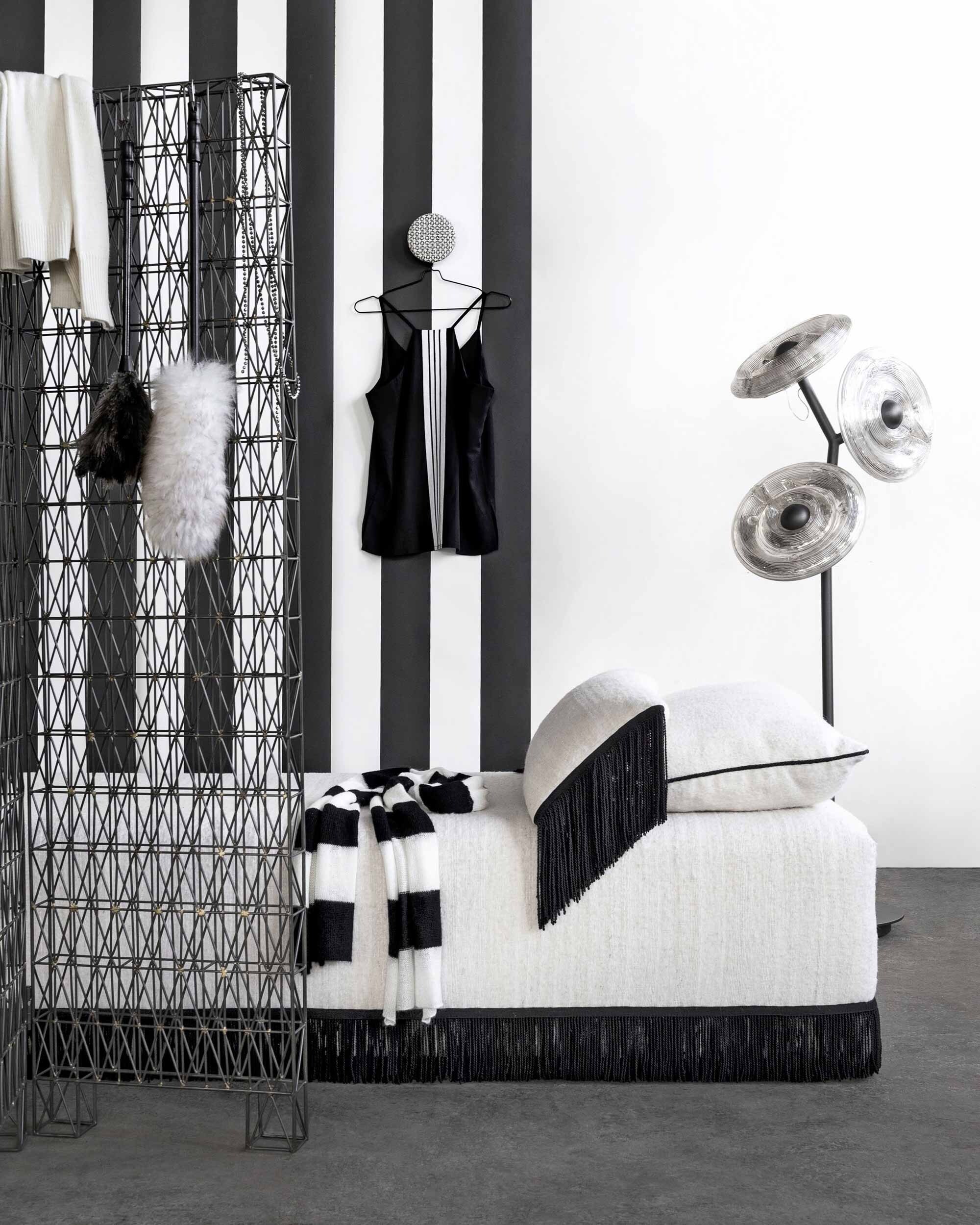 Zwart-wit gestreept behang Magnus Black van Sandberg (De Ru). Kamerscherm van staaldraad (Pols Potten). Divan Habiba met wollen bekleding (Ksähar). Lamp Sunflower (Dirk Vander Kooij). Hangknop van geborduurde stof en koper (Charlene Mullen). Plumeaus, set van 2 (HKliving). Kleding (10Days).