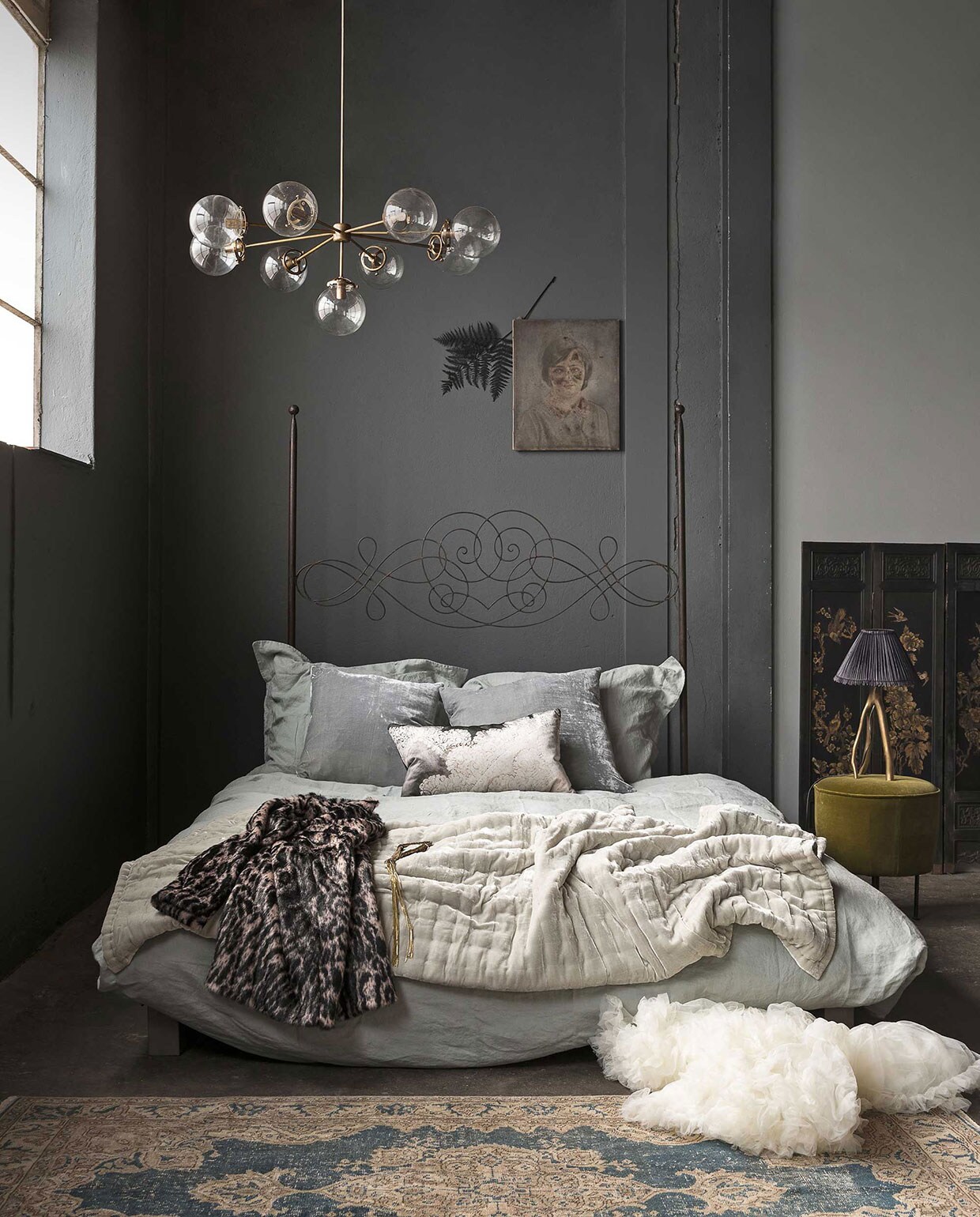 Op de muur muurverf kleur Scree van Little Greene, muurverf kleur Urban Grey van Little Greene (). Bedombouw van staal met hoog gekruld bedhoofdeind (). Linnen dekbedovertrek St. Remy inclusief twee kussenhoezen (). Kussens van zijdefluweel, groene bedsprei van zijdefluweel (). Kussen met print (). Messing kroonluchter met glazen bollen (). Schilderij meisje (). Gedroogde groenzwarte varentak (). Vloerkleed (). Drieluik kamerscherm (). Driepotige lamp met kap (). Mosgroene poef van Gubi (). Jas met dierenprint (). Petticoat (). Gouden ketting (). vtwonen 13-2017 | Fotografie Stan Koolen | Styling Marianne Luning