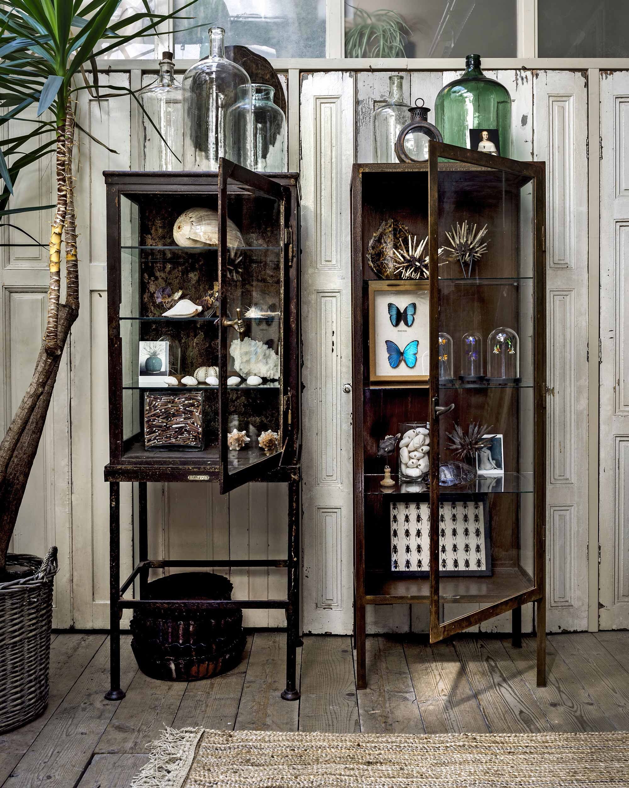 Houten panelen, vitrinekasten van staal, sisal vloerkleed, stapel dienbladen, op de kast: glaswerk, schildpadschild van kunststof op staander, oude koetslamp, in de kasten: grote schelpen, kleine schelpen, ingelijste insecten, stolpen, zwart visje op standaard (De Weldaad). Ansichtkaarten (Art Unlimited). Plantenmand privébezit.