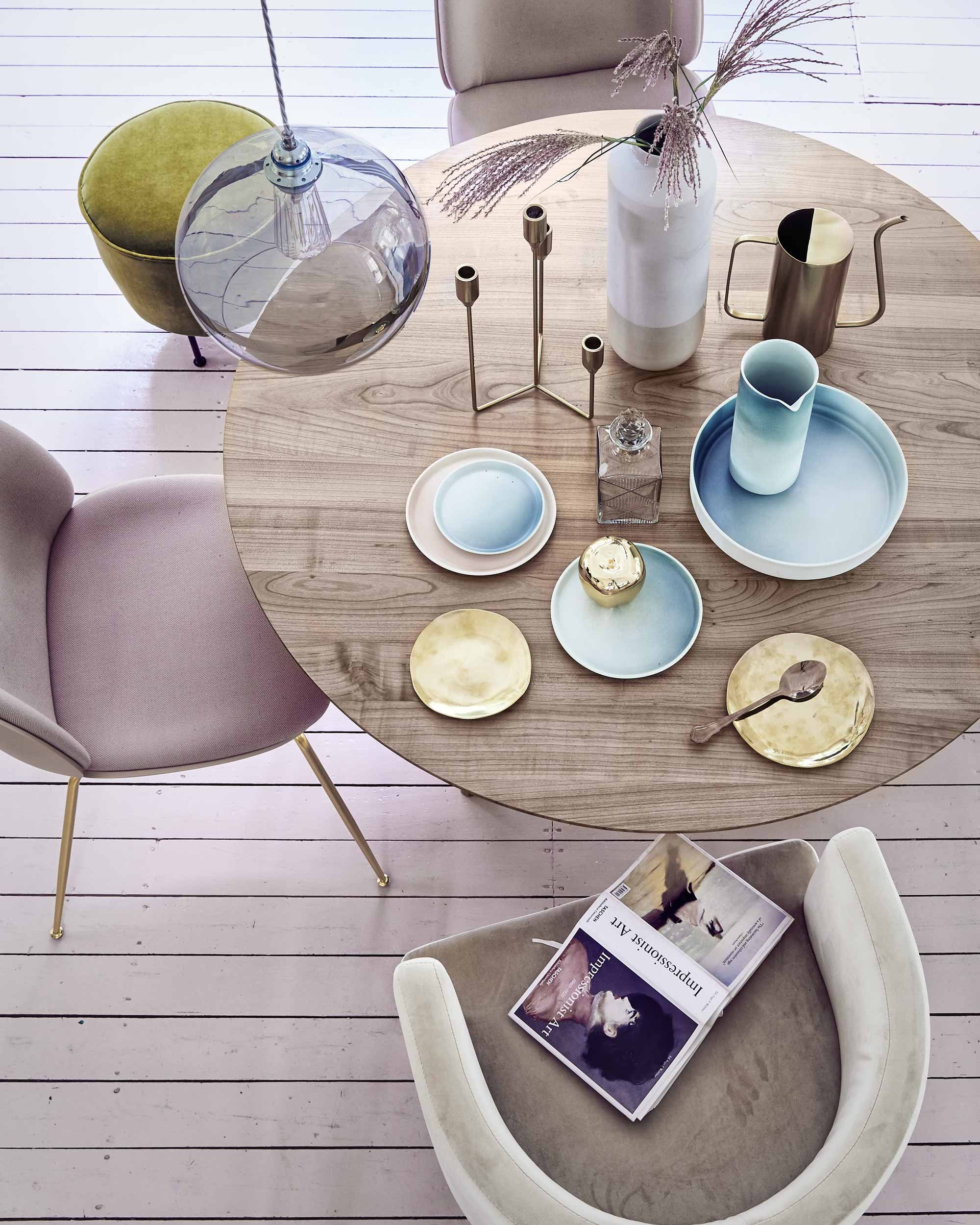 Ronde eetkamertafel, roze eetkamerstoelen van Gubi, glazen hanglamp van Ebb &amp; Flow (). Nudekleurige fluwelen fauteuil, gouden kandelaar, whiskyfles, messing gieter (). Koperkleurig bestek 12-delig (). Gouden borden, klein en groot (). Grote zachtblauwe schaal, poederroze en blauw porseleinen bord, lichtblauw bordje, grote porseleinen vaas, karaf van blauw porselein (). Kleine mosgroene poef van Gubi ().