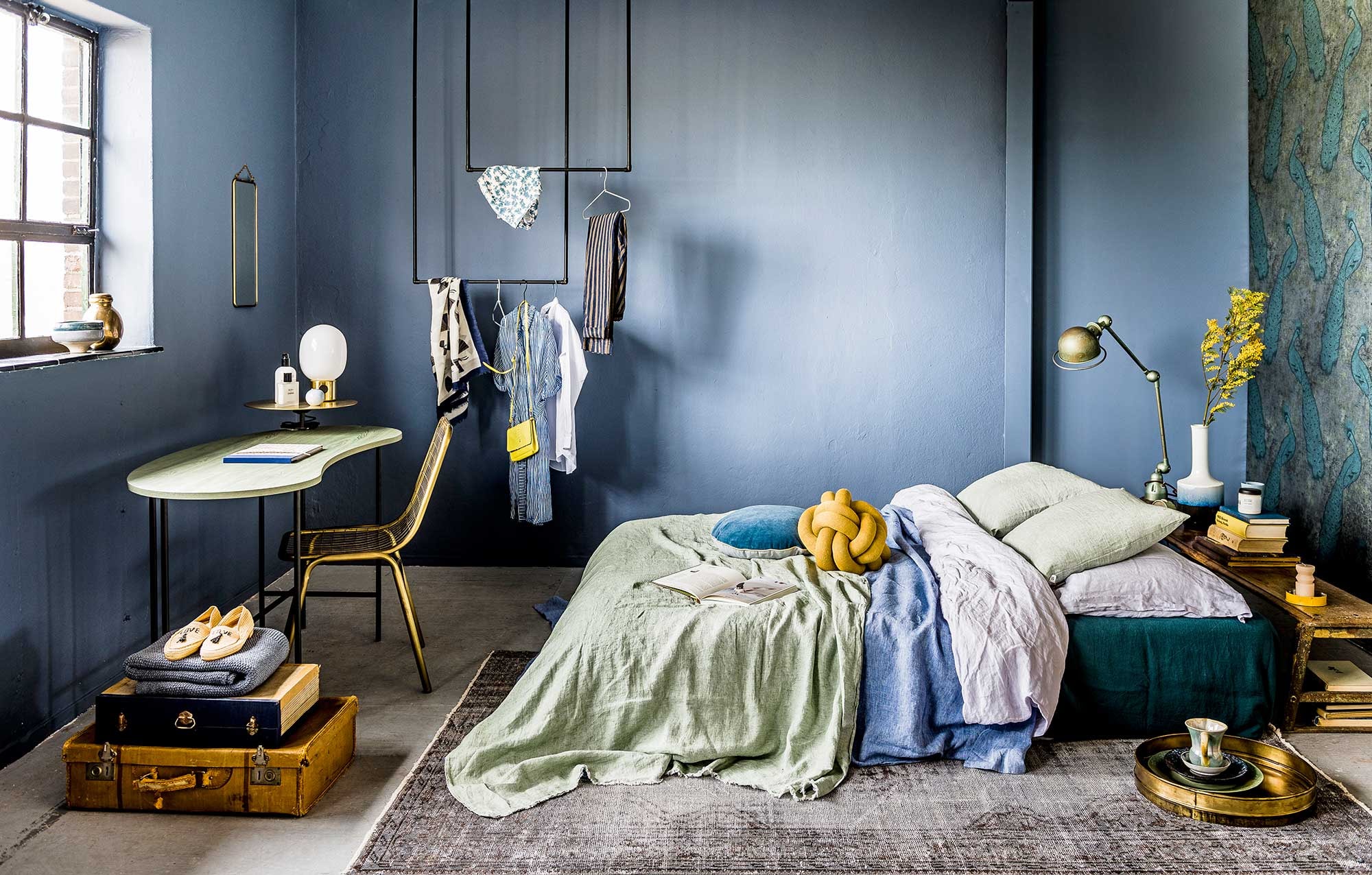 Juniper Ash 115, absolute matt emulsion (Little Greene). Twee matrassen (IKEA). Linnen donkergroen dekbedovertrek als onderlaken, denimblauw dekbedovertrek (Sissy-Boy). Stonewashed dekbedovertrek (Puur Linnen). Jadegroene plaid van By Mölle, velours rond kussen met gouden piping van Bloomingville, wit-blauwe tie-dye-vaas van Lifestyle, tafellamp op goudkeurige voet van Menu, messing draadstoel (via vtwonen). Kelim (Rozenkelim). Okergeel kussen Knot van Design House Stockholm (misterdesign.nl). Houten bankje achter het bed, spiegel koperen dienblad, borden (De Weldaad). Geel schaaltje, kaars (Six and Sons). Boeken (kringloop). Vintage Jieldé-lamp privébezit. Behang Byron van Cole &amp; Son (De Mooiste Muren). DIY-kledingrek van waterleidingbuizen (Gamma). Kledinghangers per 5 (Hay). Bureau met goudkleurige verhoging (Mobilia). Koffers (Burbri Antiek). Plaid (By Mölle). Schoenen (Love Stories). Sjaal (Six and Sons). Witte blouse, broek met streep (Sukha Amsterdam). Kan (kringloop). Beautyproducten privébezit.