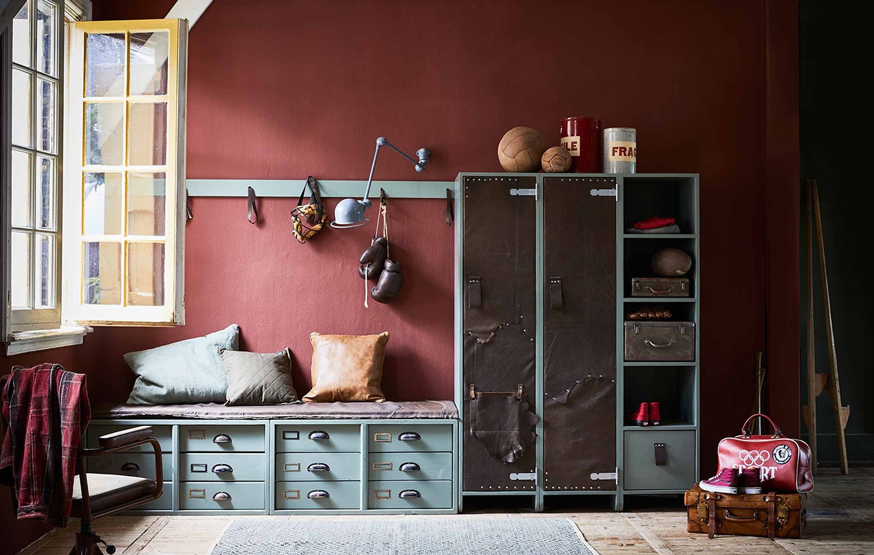 Op de wand muurverf in kleur Cardinal Red van Pure &amp; Original (). Kast met met leer beklede deuren, lage kast met lades en leren kussen, plat leren kussen, kapstok, alles op maat gemaakt (). Geverfd met lakverf in kleur Cardroom Green van Farrow &amp; Ball (). Bokshandschoenen, sporttas, metalen koffer, leren ballen, blikken Fragile, stoel, masker, American football, honkbalhandschoen (). Oude Jieldé wandlamp, leren koffer (). Leren kussen, doorgestikt legergroen kussen, groot groen kussen, vloerkleed (). Schoenen, geruit overhemd (). Stelten privébezit., vtwonen 10-2017 | Fotografie Jeroen van der Spek | Styling Cleo Scheulderman