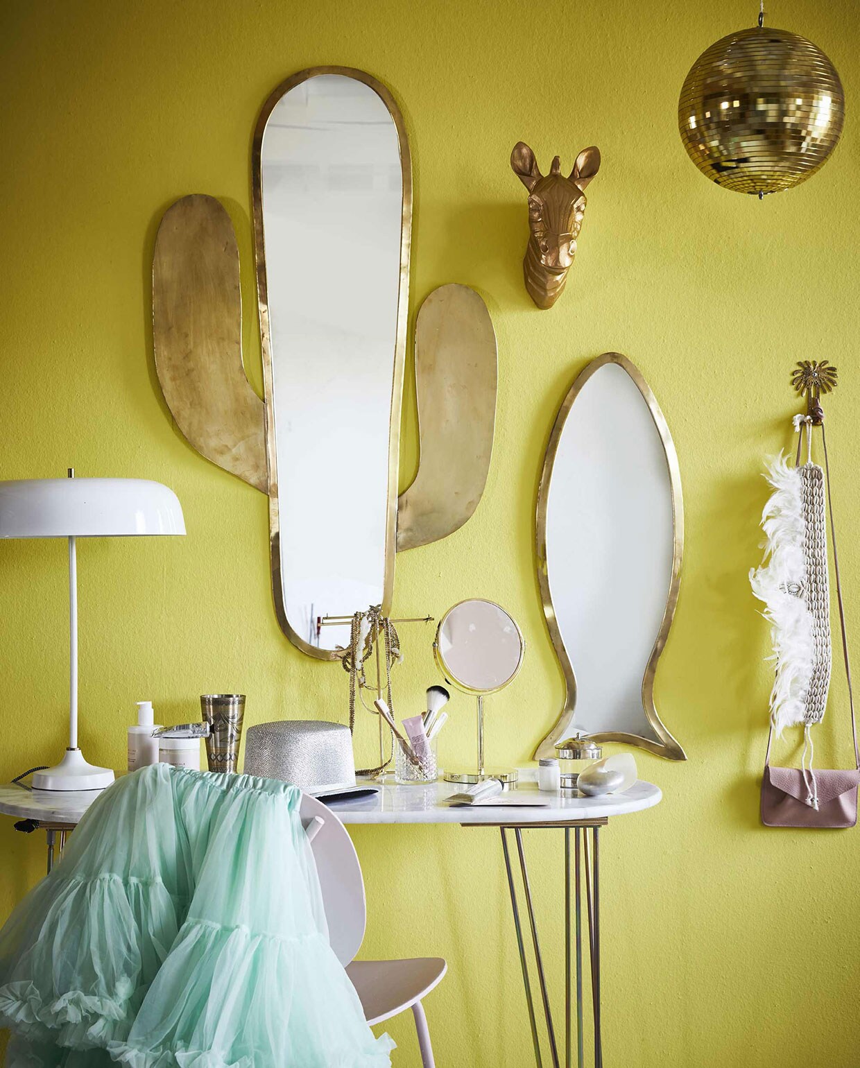 Op de wand muurverf in kleur Yellow Cake 279 van Farrow &amp; Ball (). Spiegel in cactusvorm, visspiegel (). Tafel met marmeren blad (). Witte lamp (). Verentooi met kralen, schelp, metalen beker, ketting, armband, potje met zilveren deksel, ophanghaak palmboom (). Roze tasje (). Goudkleurig zebrahoofd (). Discobal (). Petticoat (. Spiegel op voet, sieradenhouder, stoel (). vtwonen 10-2017 | Fotografie Jeroen van der Spek | Styling Cleo Scheulderman