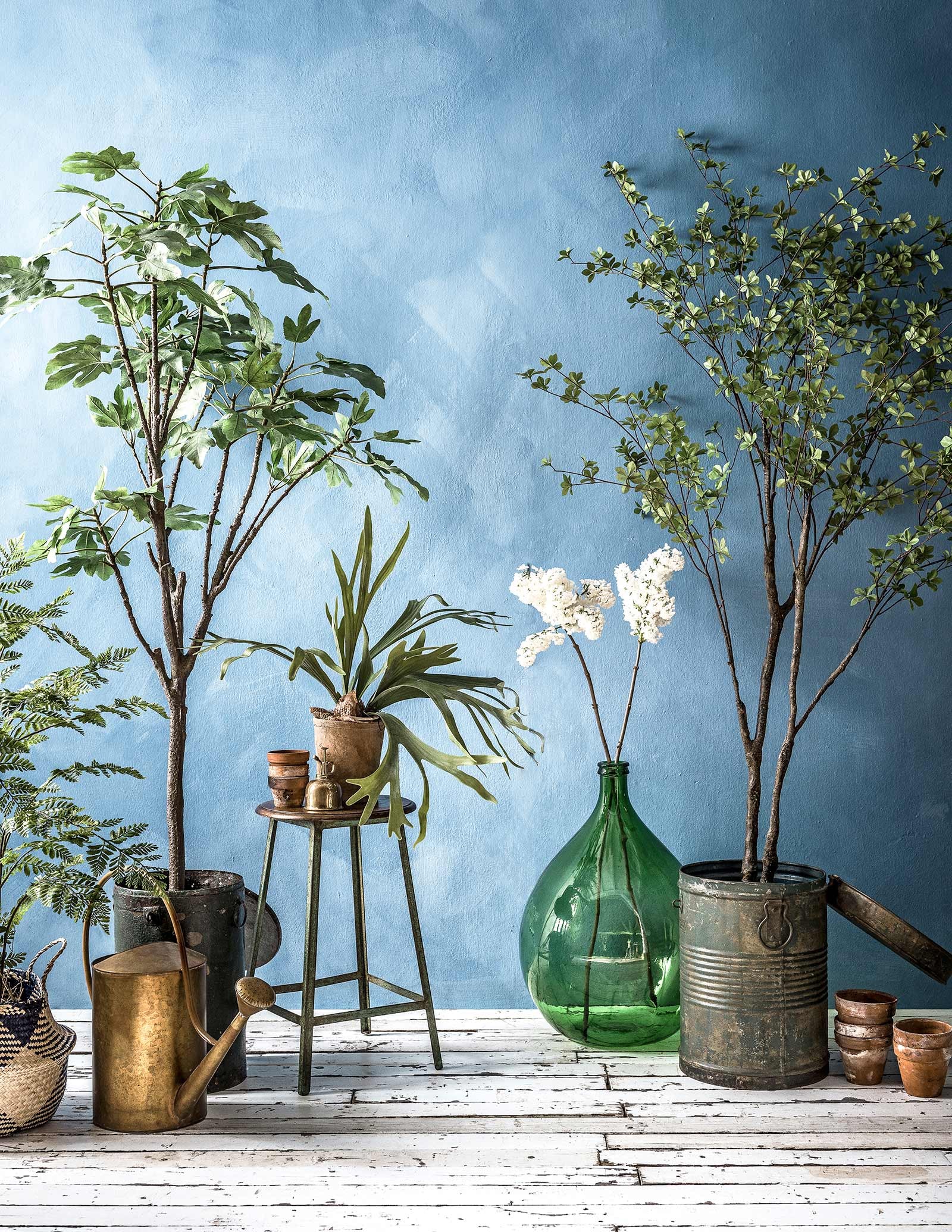 Muurverf in kleur Periwinkle van Bauwerk Color (). Gevlochten mand met blauwe vlakken (). Kleine kunstplanten in pot, grote kunstplanten en -boompjes, groene metalen ton met deksel, houten kruk met metalen onderstel (). Messing gieter (). Oude terracotta bloempotten, witte seringen () Messing plantenspuit (). Groene glazen flesvaas ().