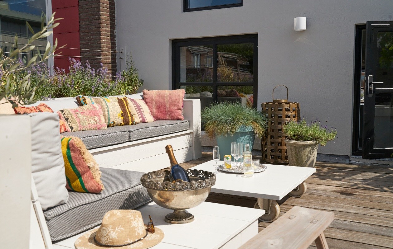 Loungemeubel en tafel op wielen eigen ontwerp. Kruk en geweven lantaarn Loods 5. Koperen buitendouche Seletti. Kussens op de bank Tim’s Kussens, kleurige kussens Bohemian du Maroc. Zilveren schaal op voet ooit bij de Bijenkorf gekocht. vtwonen 08-2020 | styling Wilma Custers | fotografie Luuk Geertsen